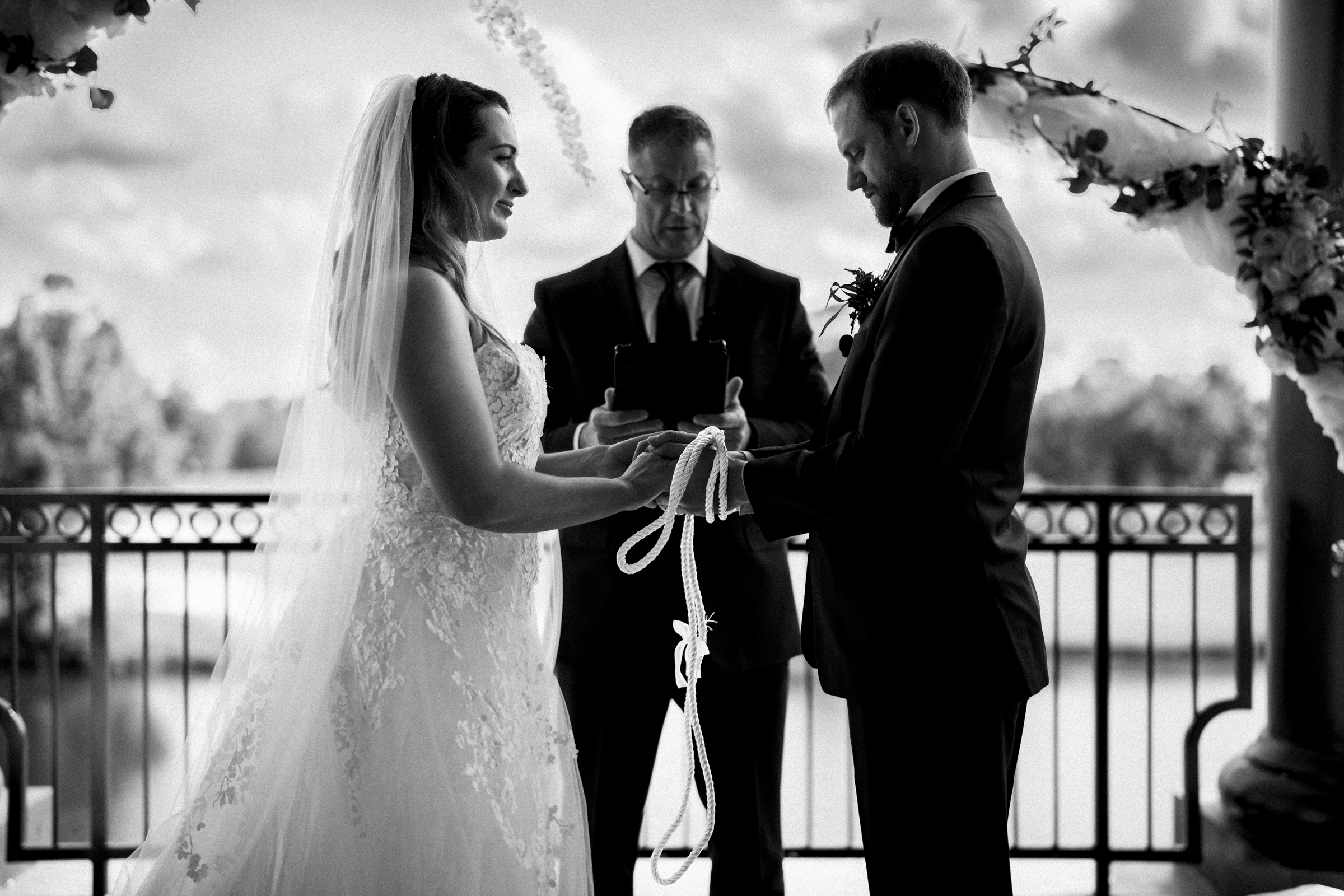 A bride and groom stand facing each other, holding hands with a cord, during a beautiful river landing wedding. The officiant stands behind them, book in hand. The background features a serene landscape with a railing and trees.