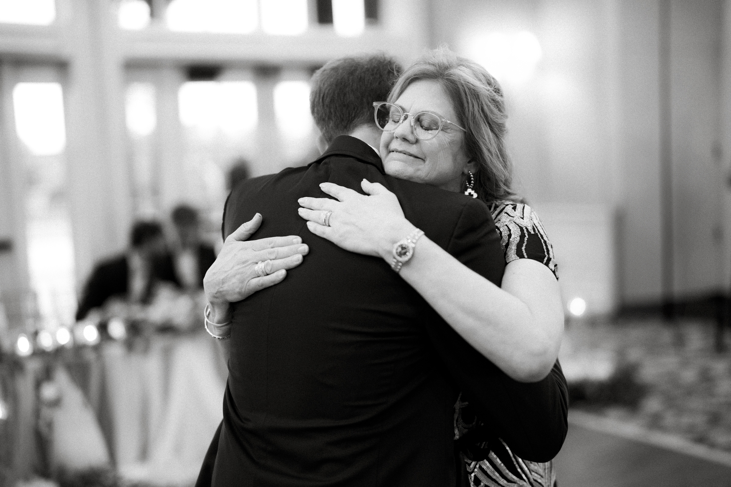 A woman with glasses and a patterned dress warmly hugs a man in a suit at a formal river landing wedding. They both appear content in the black and white photo, with a softly blurred background of the reception area.