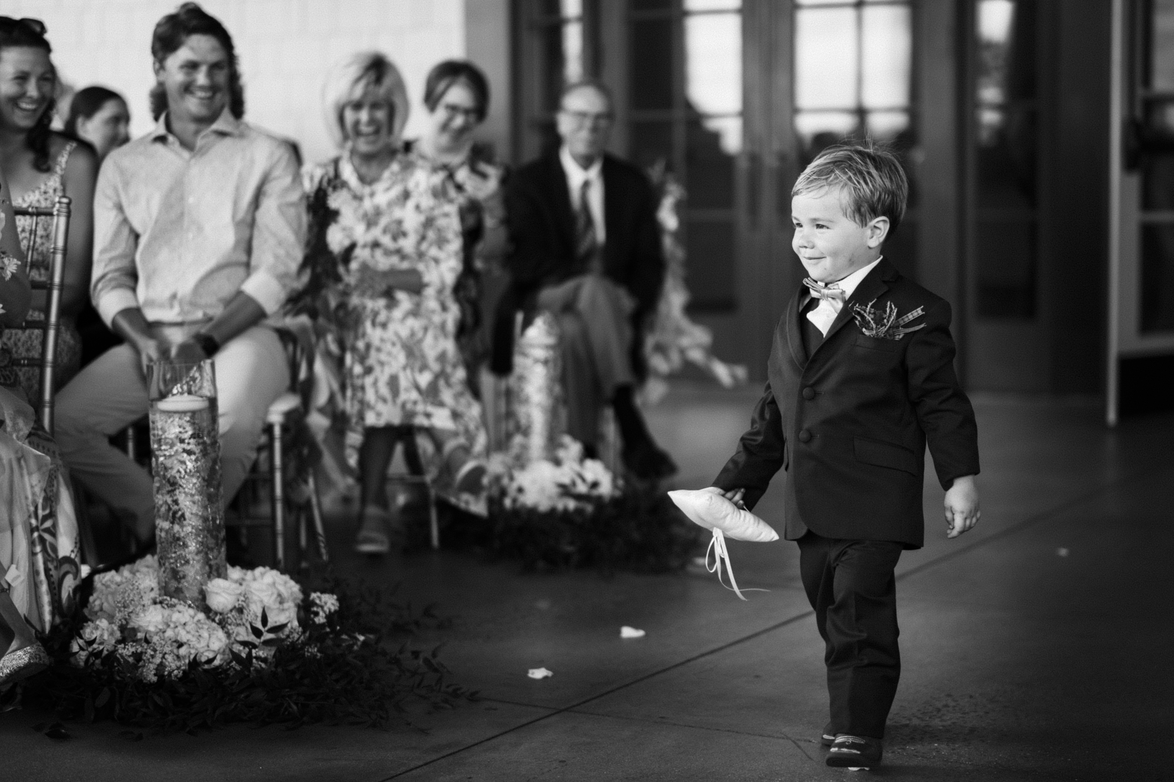 A young boy in a suit walks down an aisle at the river landing wedding, carrying a pillow and smiling. Guests are seated on either side, watching him. The scene unfolds in elegant black and white.