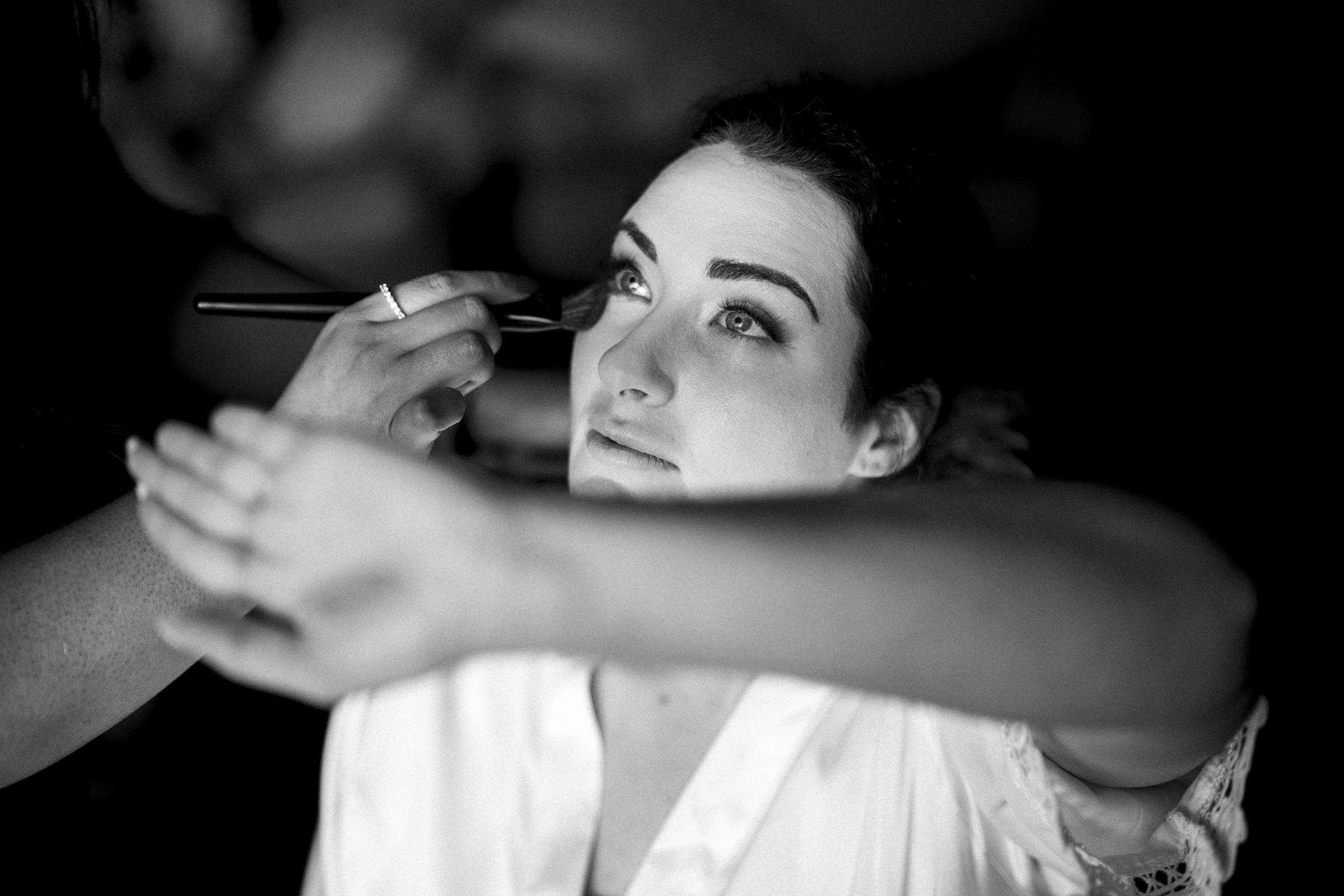 A woman in a white robe, preparing for her River Landing wedding, has her makeup done with a gentle brush. Her arm is raised, partially covering her eyes. The black and white image beautifully captures her serene expression.