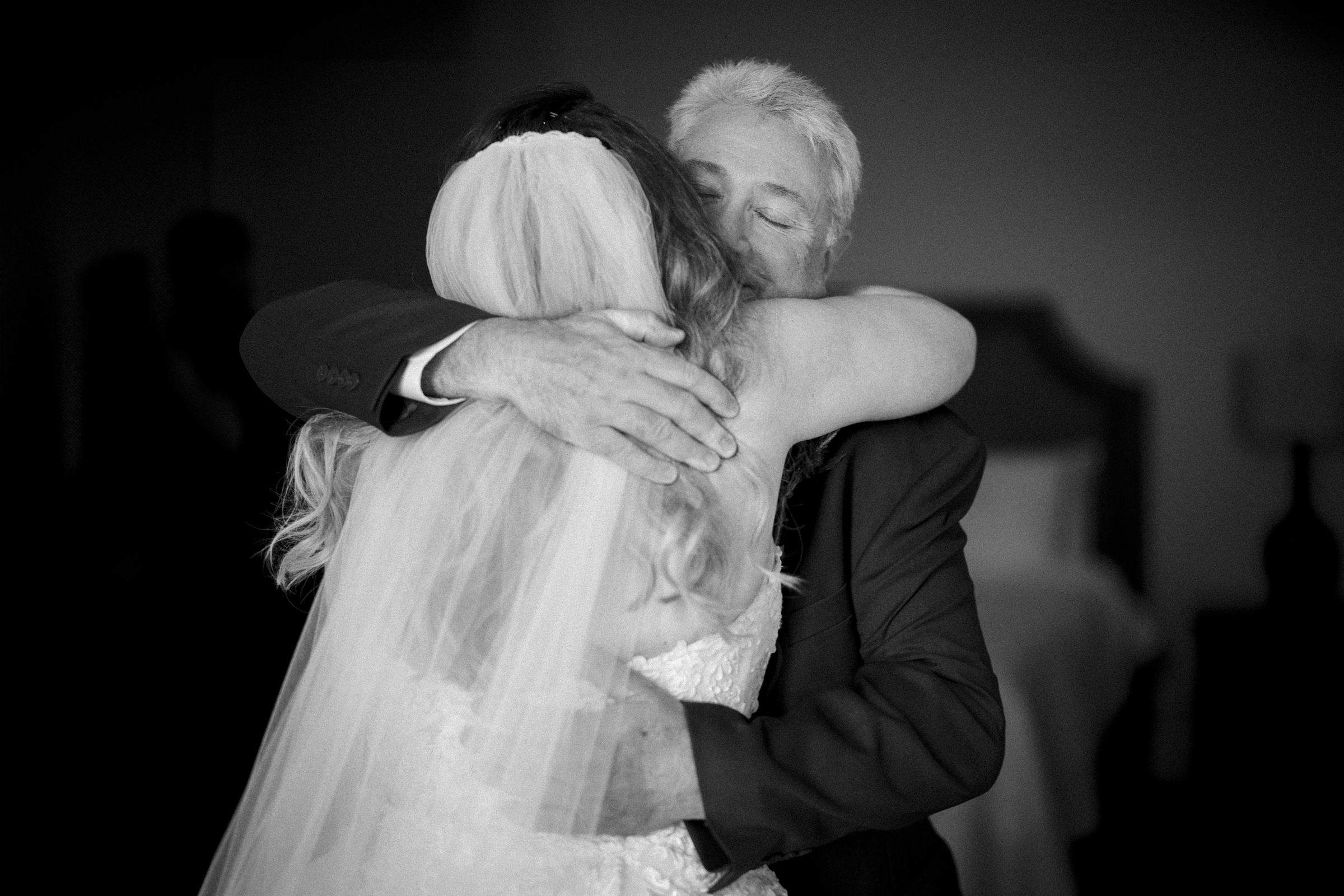 A black and white photo captures a tender embrace between a bride in a veil and strapless gown and an older man in a suit. The softly blurred background hints at an intimate river landing wedding setting.