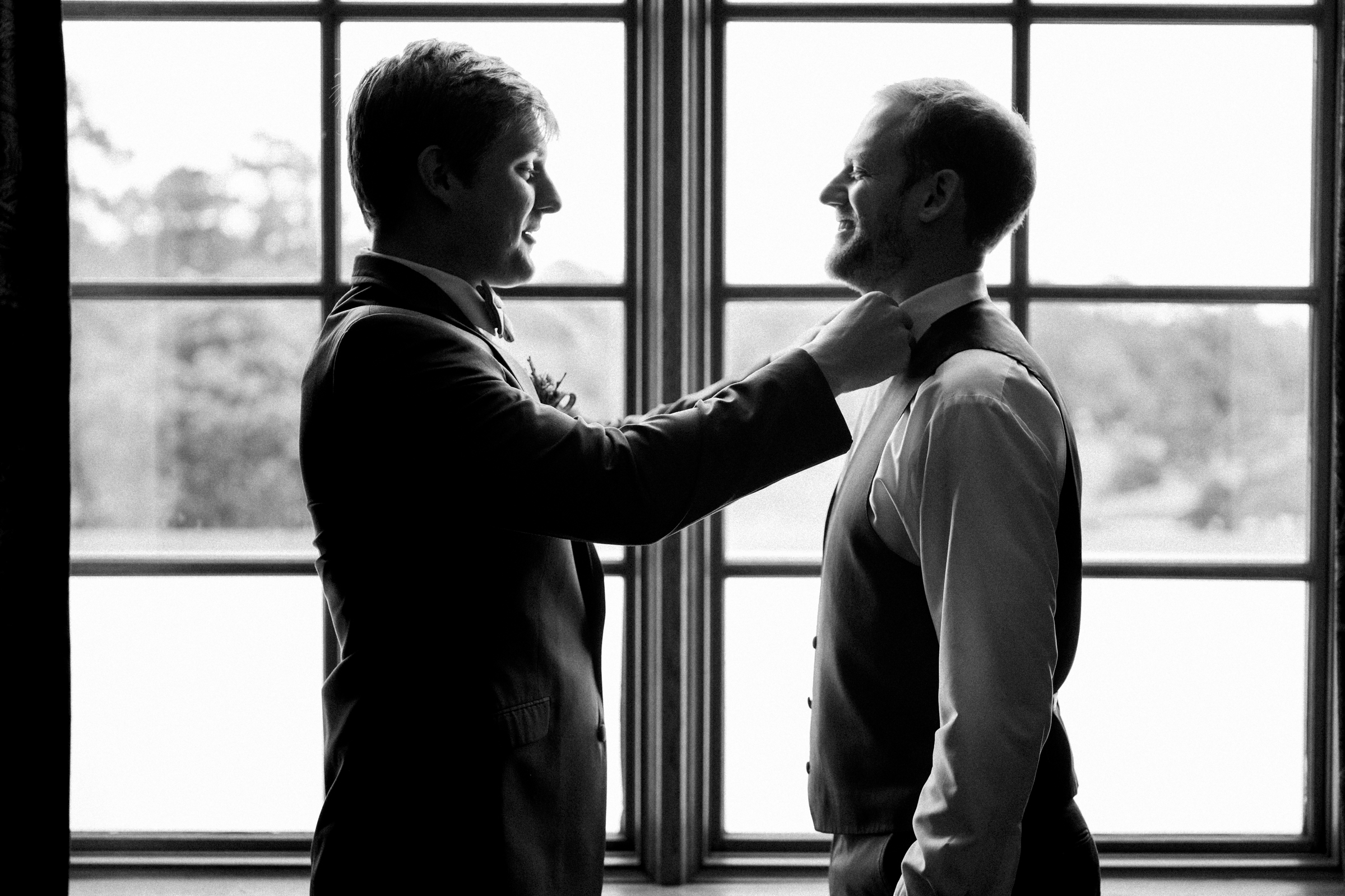 Two men in formal attire stand facing each other in front of a large window at River Landing. One is adjusting the other's tie, both smiling. The black and white scene conveys a moment of friendship or preparation for a wedding event.