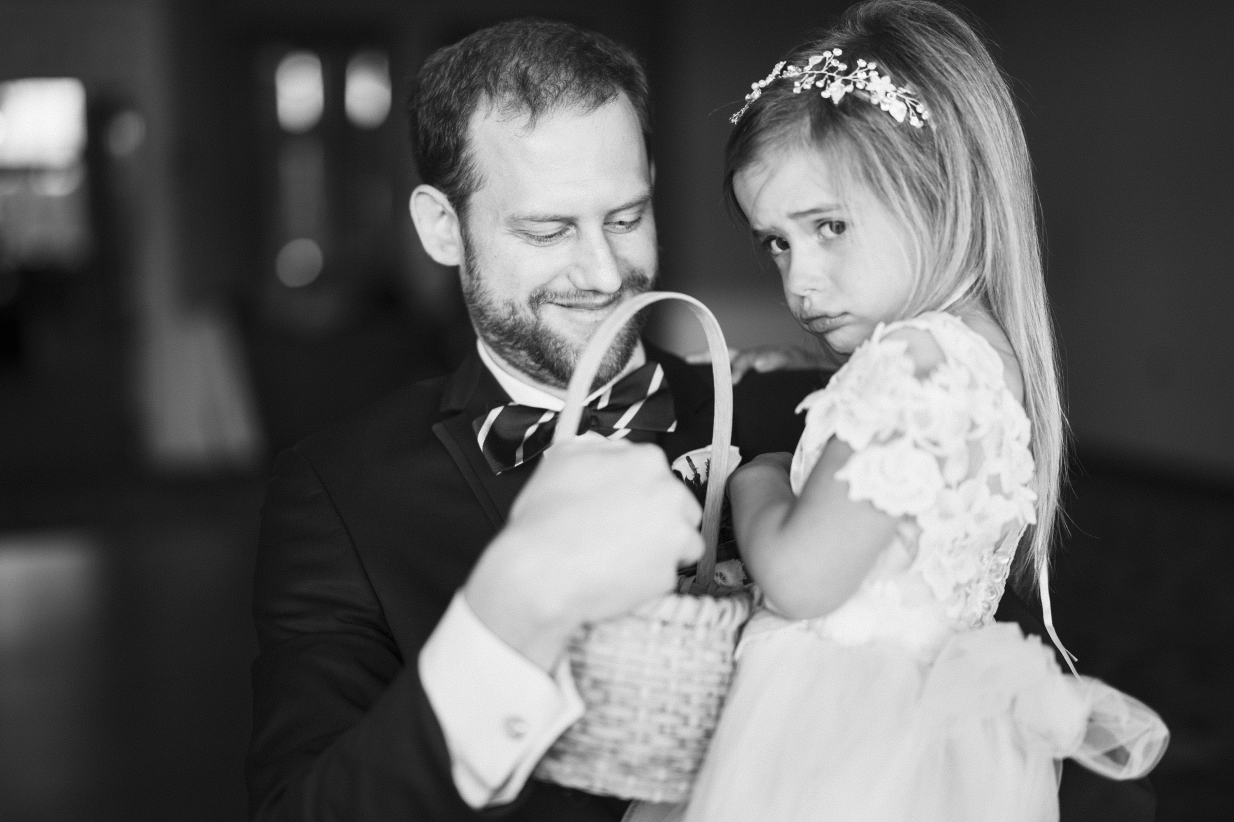 A man in formal attire holds a young girl dressed in a fancy dress with a small tiara, likely at a river landing wedding. She clutches a wicker basket and looks slightly pouty, set against an indoor scene with a soft-focus background.