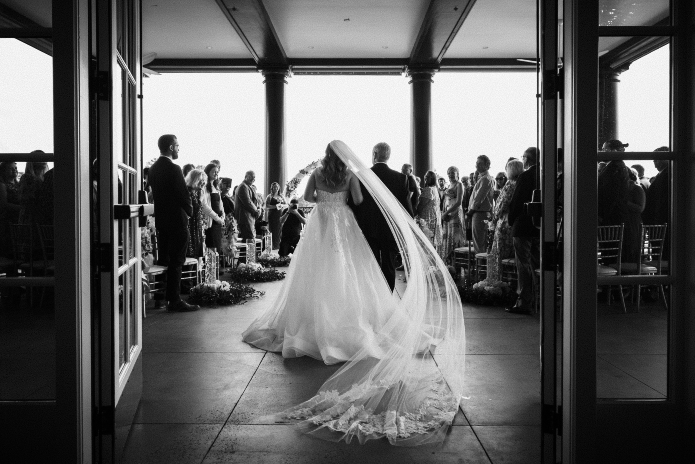 A bride in a flowing gown and veil walks down the aisle with an older man, likely her father. Guests at the River Landing wedding stand on either side in an elegant venue, capturing the moment in black and white.