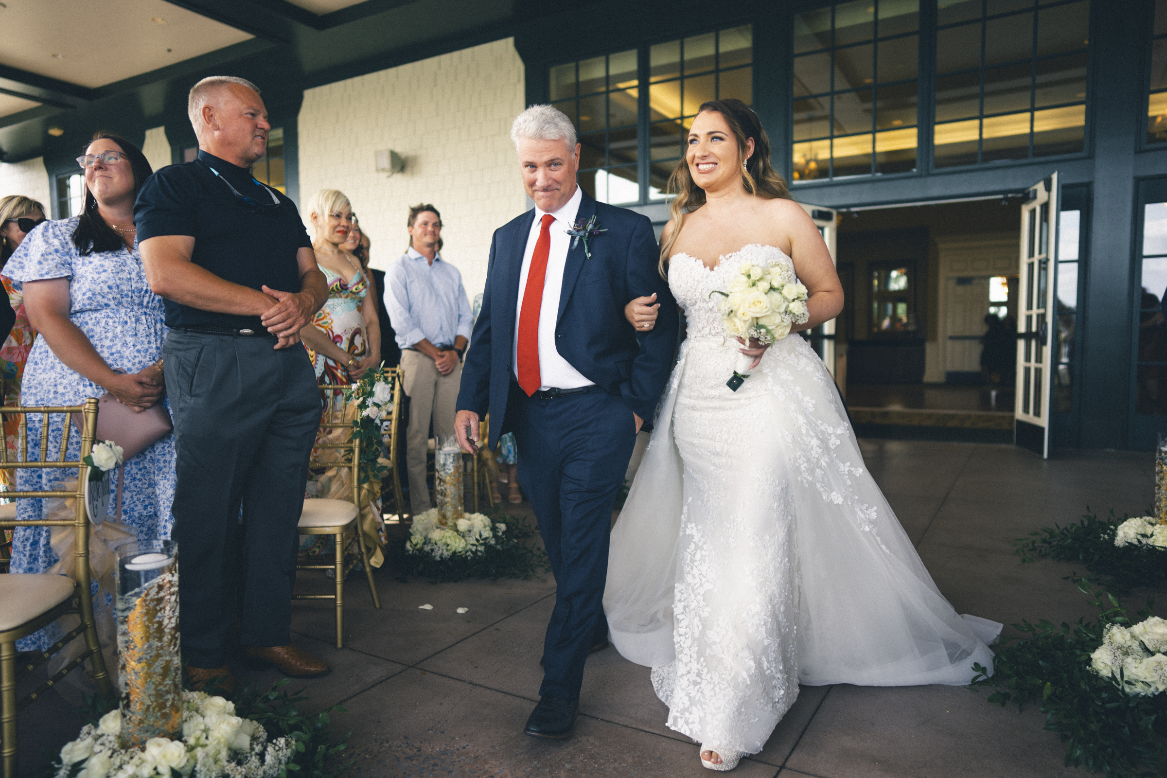 At a beautifully decorated indoor venue, a bride in a white gown walks down the aisle at a stunning river landing wedding. Accompanied by an older man in a suit and red tie, she beams as she carries her bouquet past seated guests. The flower-adorned aisle adds to the enchanting scene.