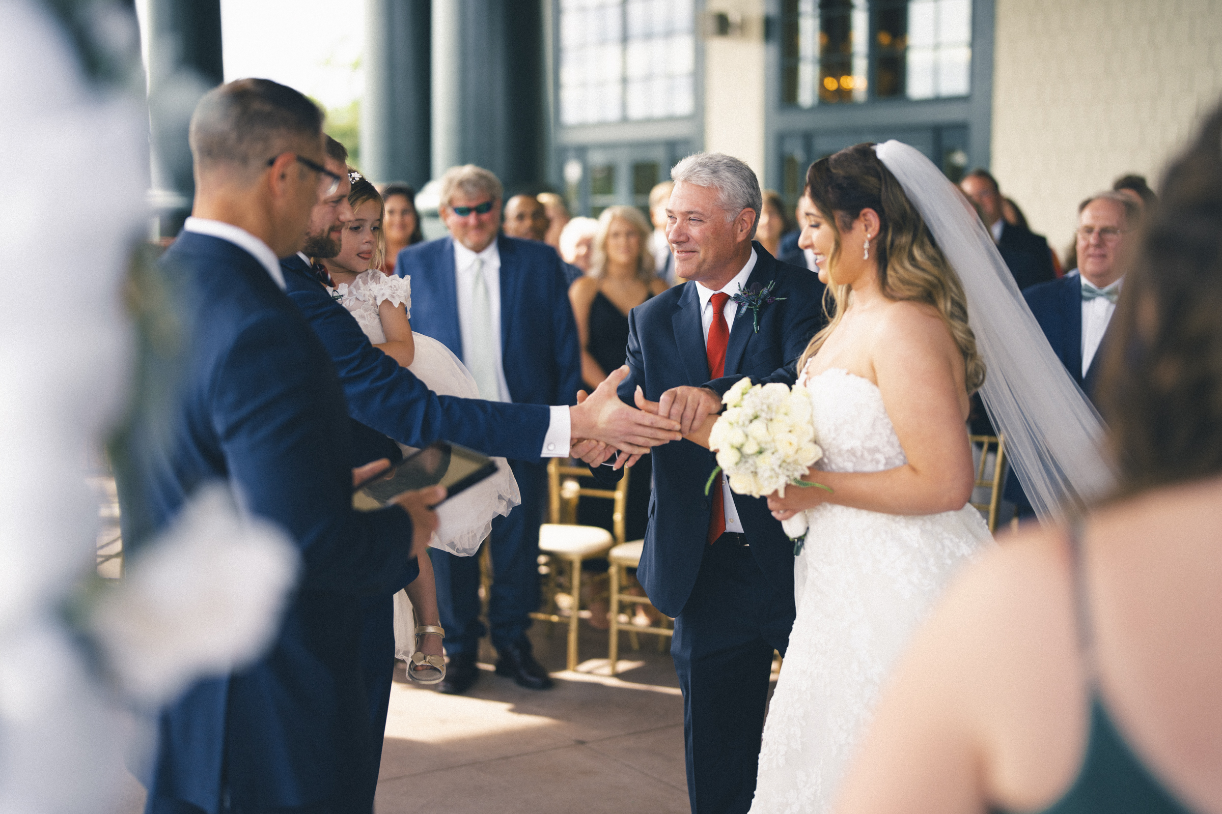 A bride in a white gown and veil smiles as an older man in a suit escorts her at the river landing, shaking hands with the groom. Guests watch, some standing and some seated. The outdoor setting features a bright backdrop and floral decorations, perfect for this charming wedding celebration.