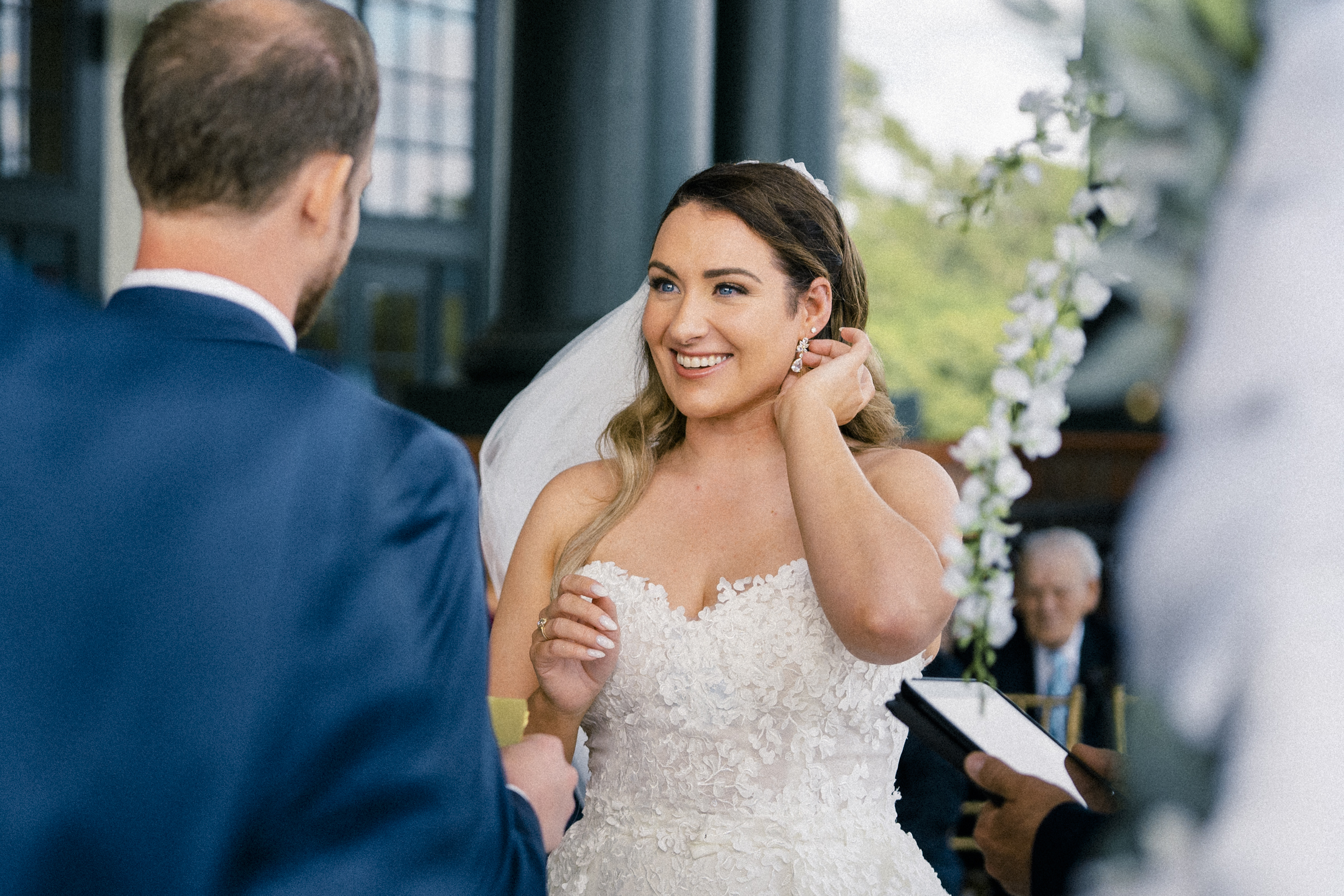 A smiling bride in a white, floral-textured wedding dress stands next to a groom in a blue suit, adjusting her earring. They are at an enchanting river landing wedding ceremony, with a floral arch nearby and guests seated against the scenic backdrop.