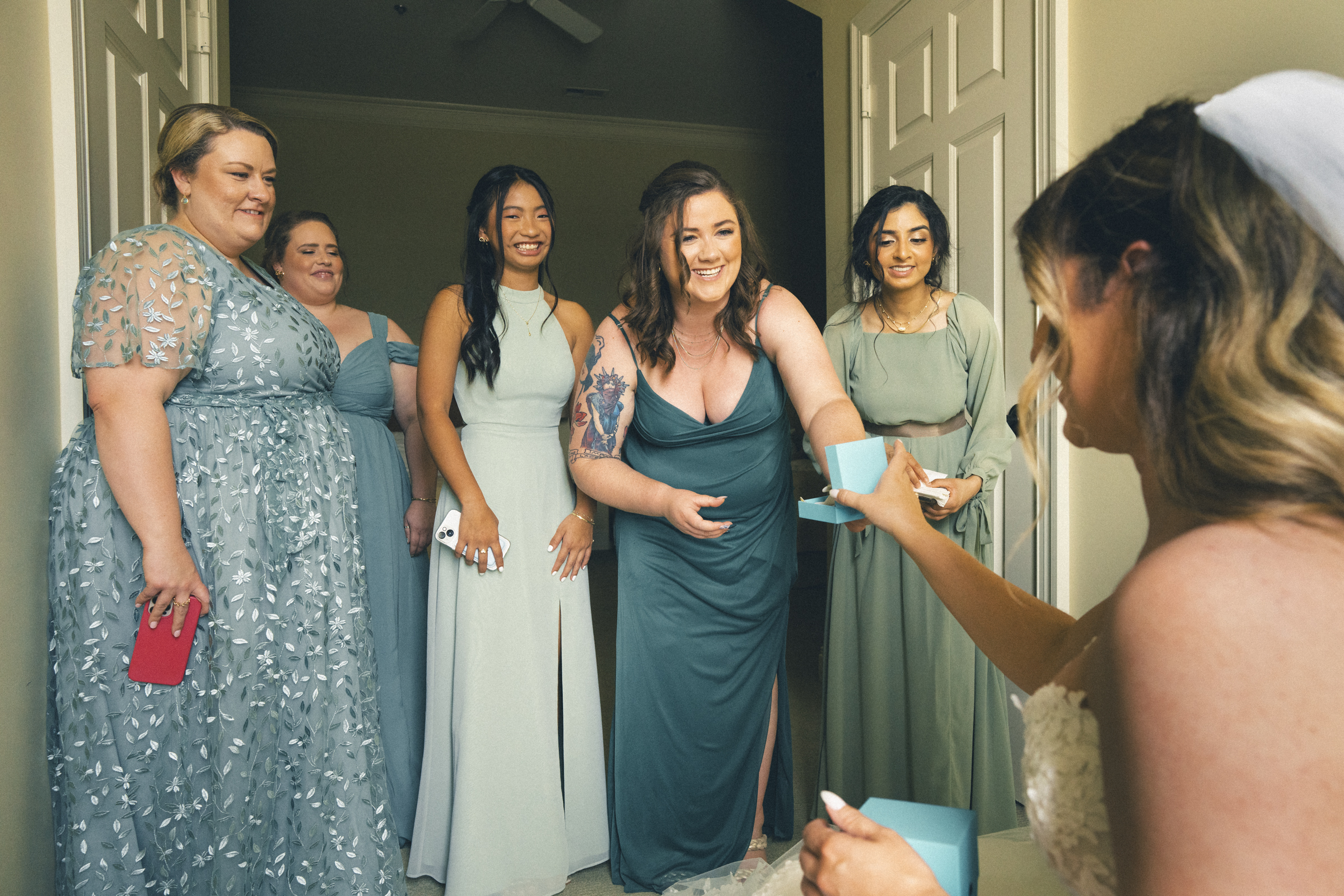 In a warmly lit room at River Landing, a bride smiles as she reaches for a blue gift box from her five bridesmaids. Each wears a unique shade of green, adding to the joyful ambiance of this special wedding moment.