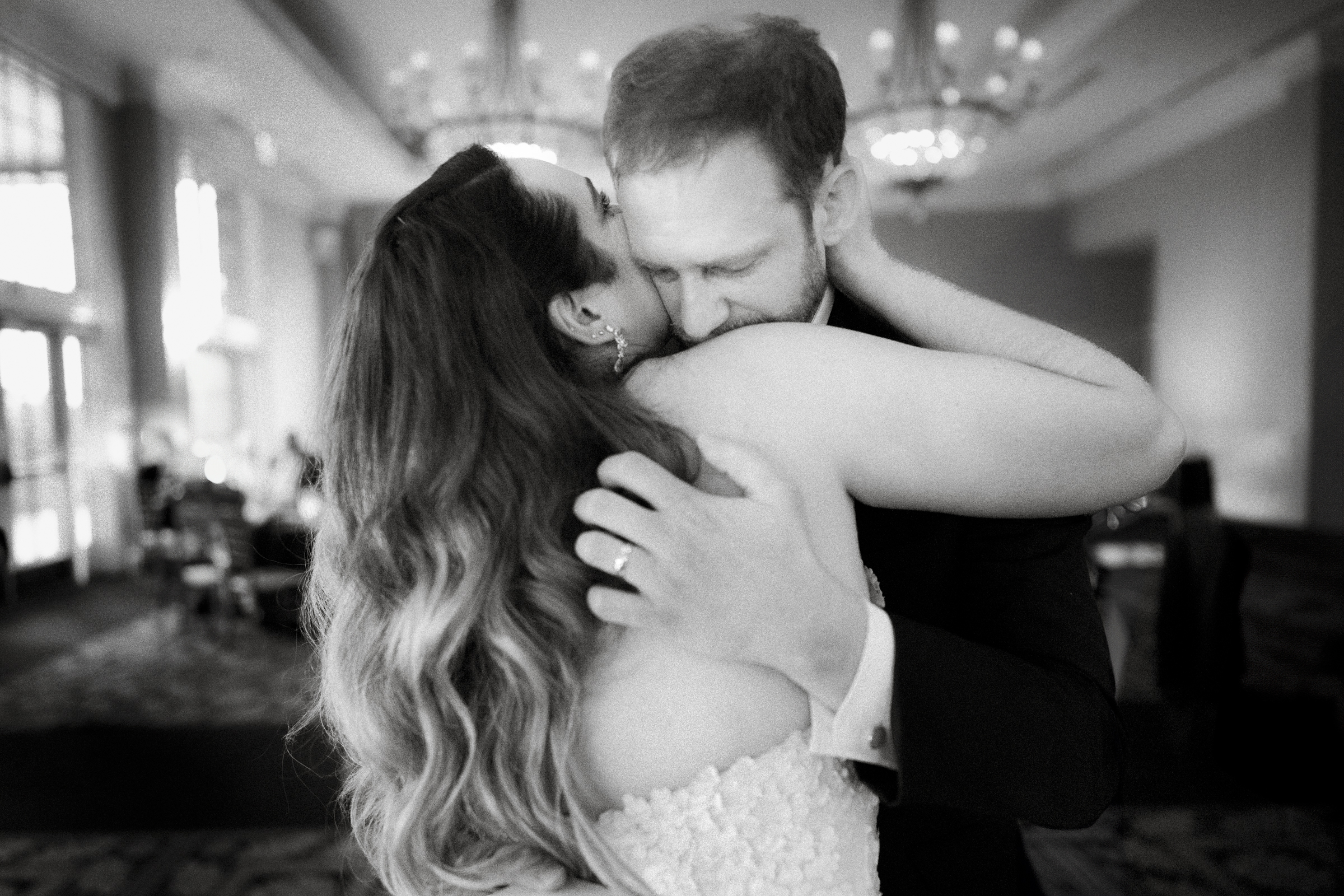 A black and white photo captures a couple dancing closely at a River Landing wedding. The woman, with long wavy hair, smiles with her face partly hidden, while the man embraces her warmly. The softly lit ballroom, adorned with chandeliers, enhances the romantic ambiance.