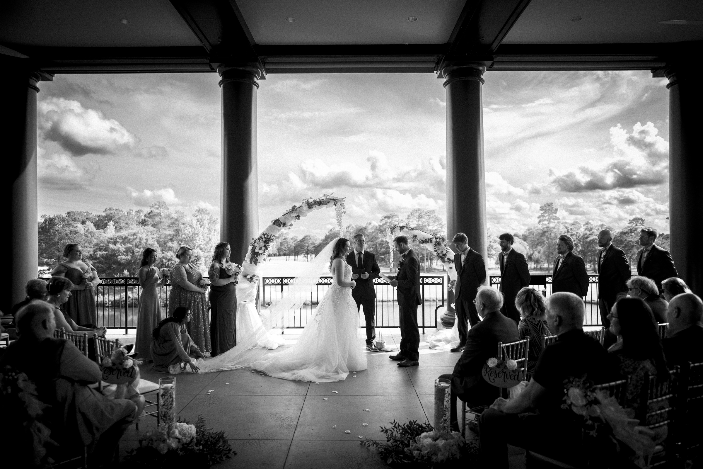 A black and white photo captures a river landing wedding ceremony on a terrace. The bride and groom stand under a floral arch, embraced by their bridal party and guests. In the background, lush trees frame the scene against a cloudy sky.