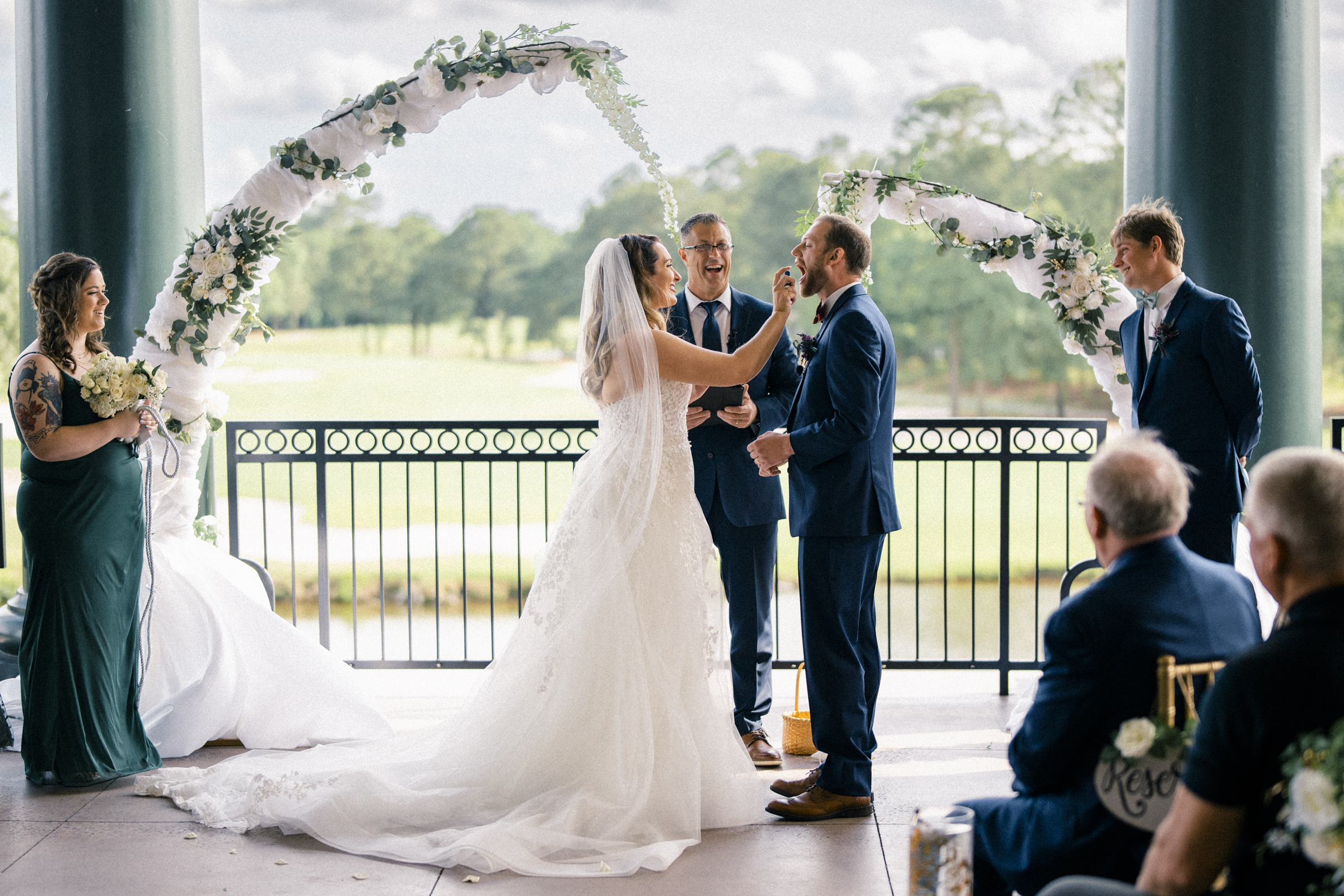 A bride and groom stand under a floral arch during their river landing wedding ceremony. The bride, in a white gown, gently touches the groom's face. Guests and a minister observe, while the bridesmaid holds a bouquet. The idyllic setting is outdoors with lush greenery in the background.