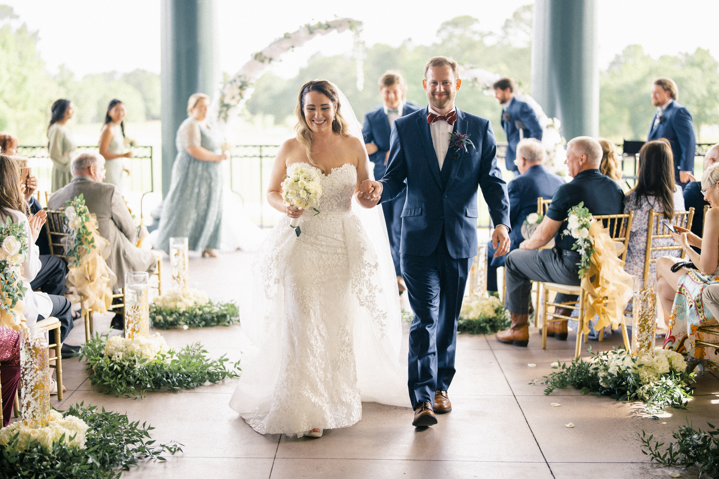 A bride in a white lace gown and a groom in a navy suit hold hands, smiling as they walk down the aisle at their river landing wedding. Guests seated on either side, surrounded by lush greenery and floral decorations, admire the arch adorned with white flowers in the background.