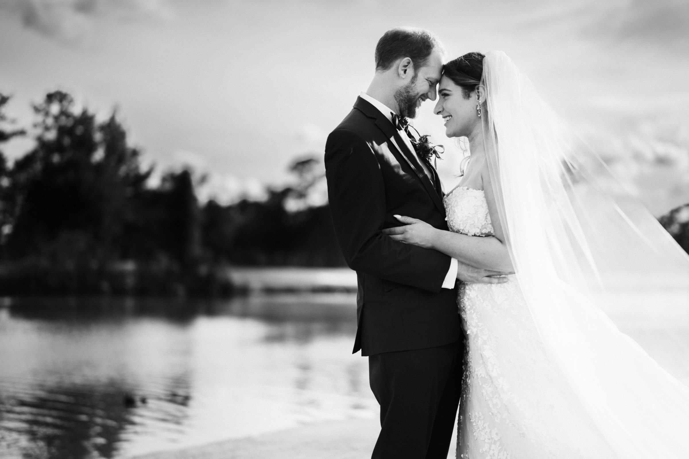 A bride and groom embrace beside a serene river landing. The bride wears a long veil and a lace dress, while the groom is in a dark suit. They gaze at each other affectionately, surrounded by trees and calm waters under a clear sky, creating the perfect wedding portrait.