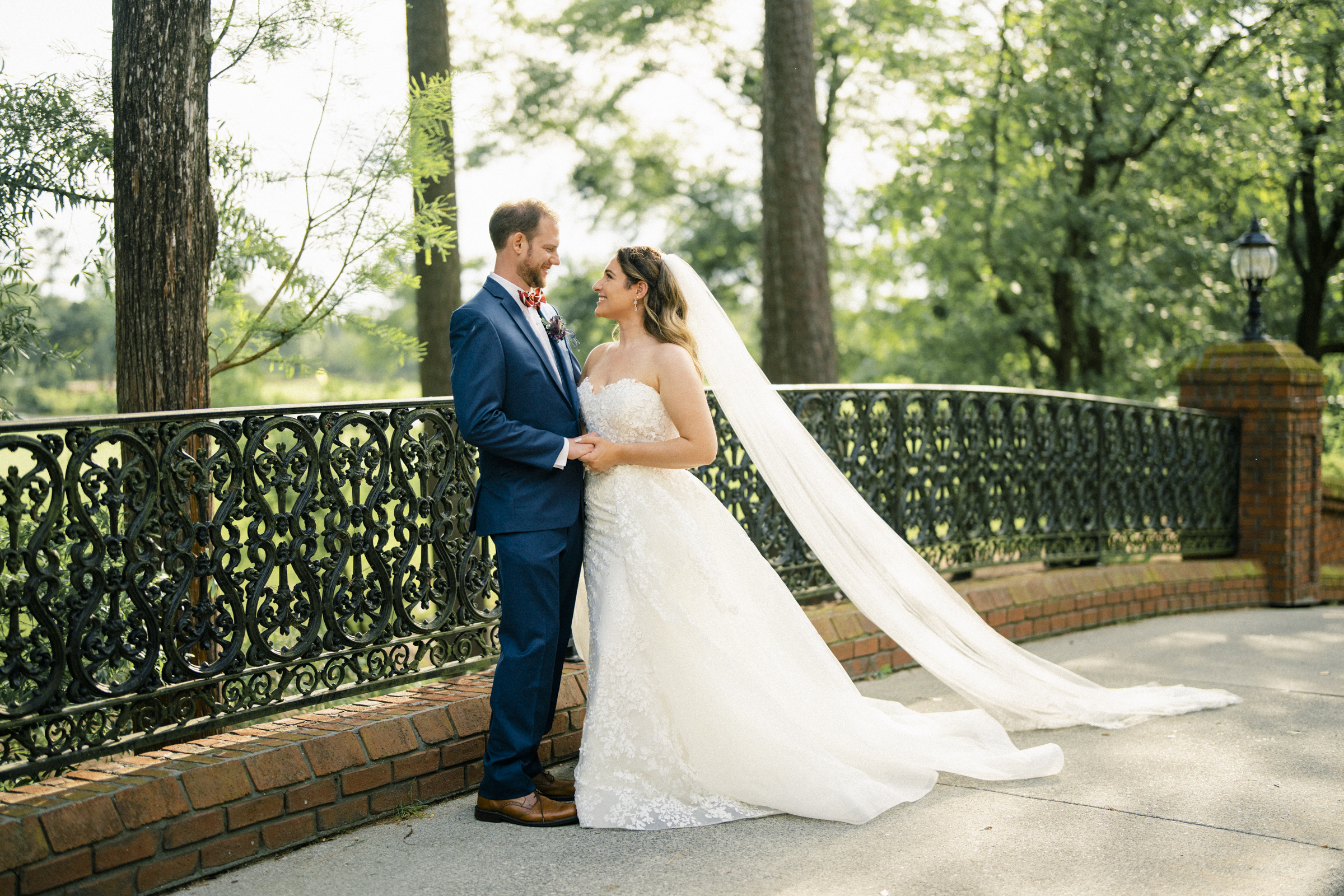 A bride and groom hold hands, gazing at each other on a sunlit path at River Landing. The bride wears a flowing white dress with a long veil, while the groom is in a blue suit. They stand in front of a decorative wrought-iron fence with trees in the background.