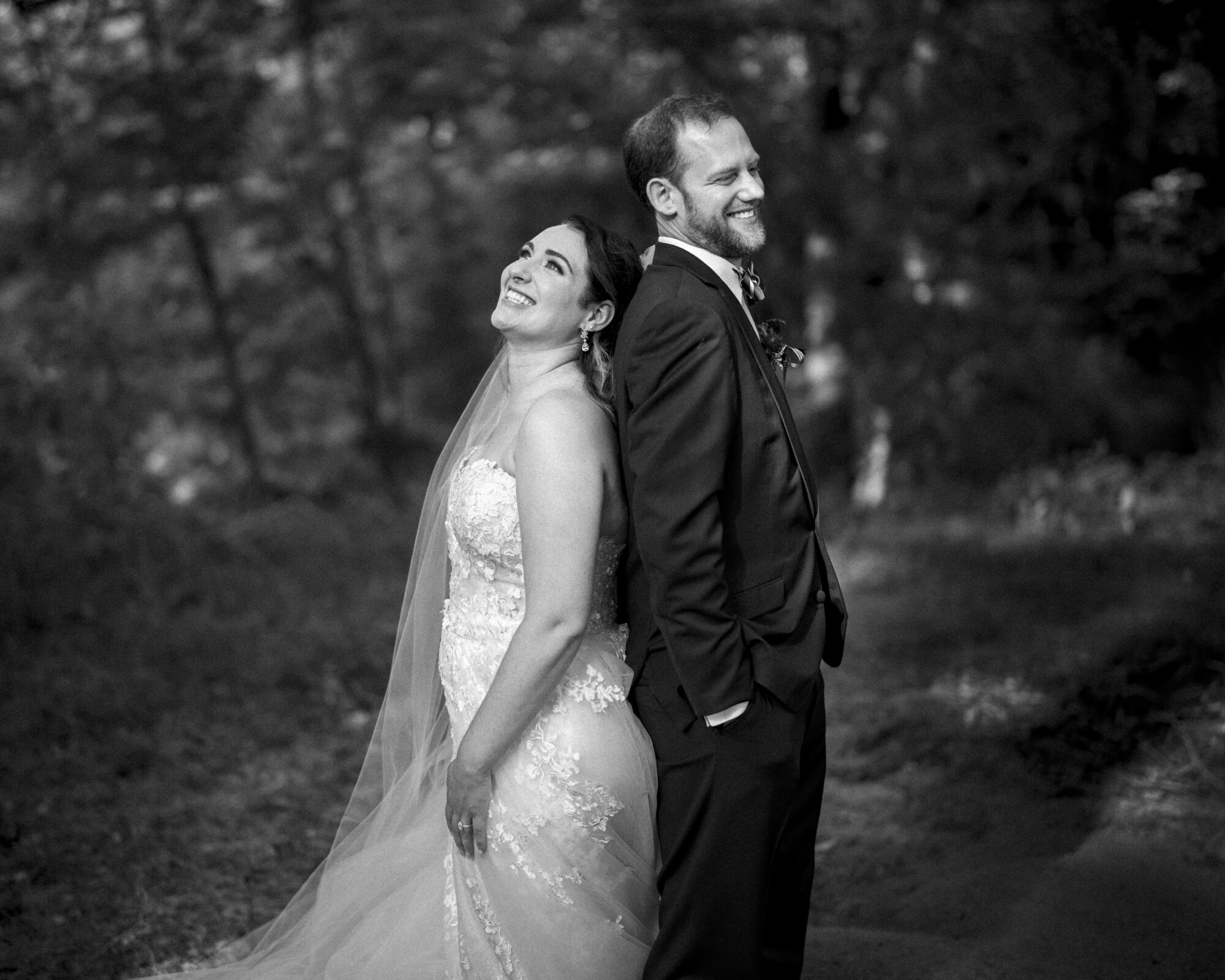 A bride and groom stand back-to-back, smiling and laughing by the river landing in a wooded outdoor setting. The bride wears a lace gown with a long veil, while the groom is in a dark suit and tie. Shadows of trees create a textured background.