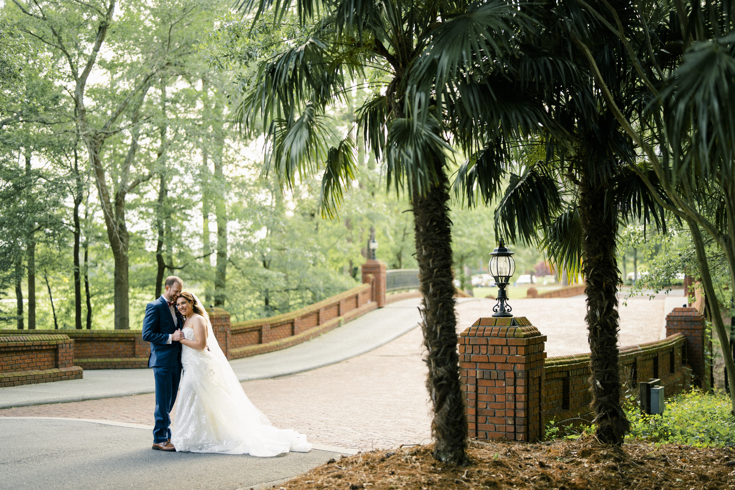 A bride and groom embrace on a tree-lined brick path near a decorative lamp post, perfectly capturing their river landing wedding. The bride wears a flowing white gown, the groom a blue suit. The scene is peaceful, surrounded by lush greenery and tall palm trees.