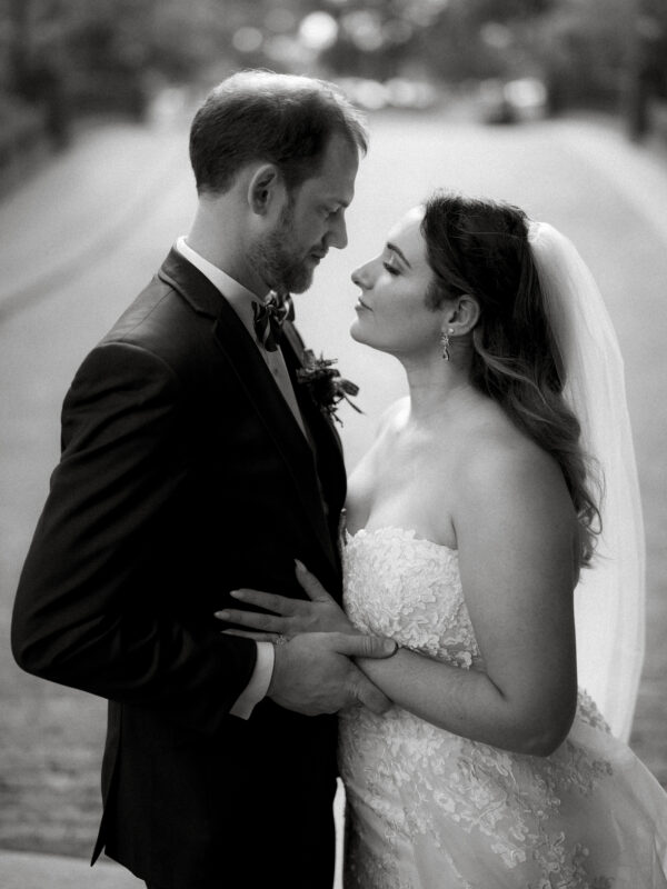 A black and white photo captures a tender moment between a bride in a strapless lace gown and veil, and her groom, who sports a bow tie. They stand closely, hands entwined, at the enchanting River Landing wedding venue, with trees lining the path in the background.