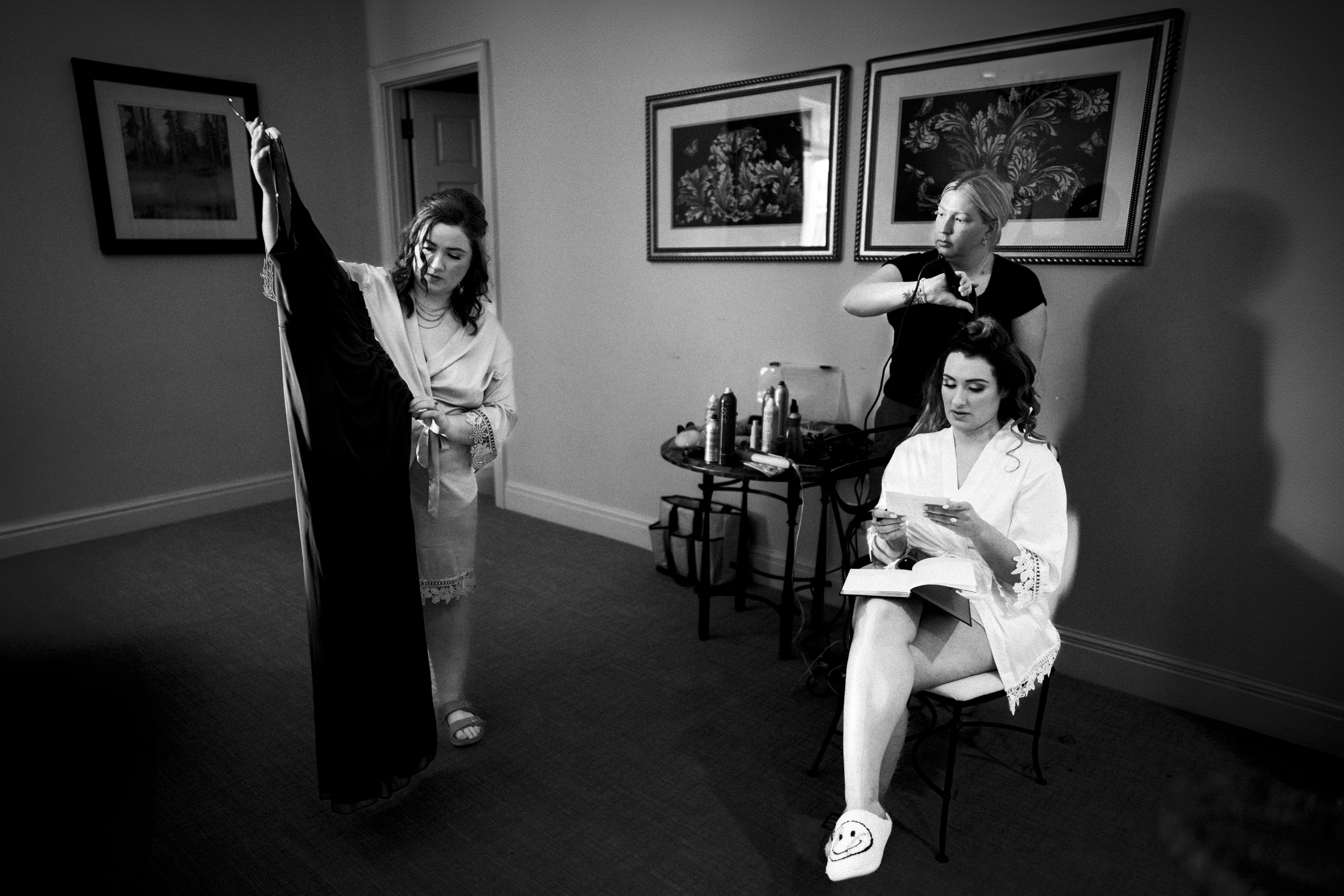 A black-and-white image captures a wedding preparation scene: a woman in a robe holds up a dress, another sits reading, and the third styles her hair. The room, adorned with hairdressing tools and framed art on the walls, exudes an air of elegance reminiscent of river landing serenity.