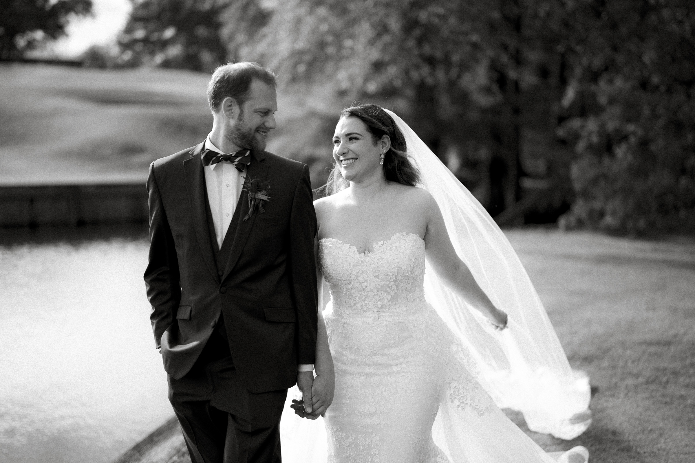 A bride and groom hold hands and smile at each other while walking outdoors. The bride wears a lace gown with a long veil, and the groom is dressed in a suit with a bow tie. They stroll near the river landing, with trees providing a picturesque backdrop to their perfect day.