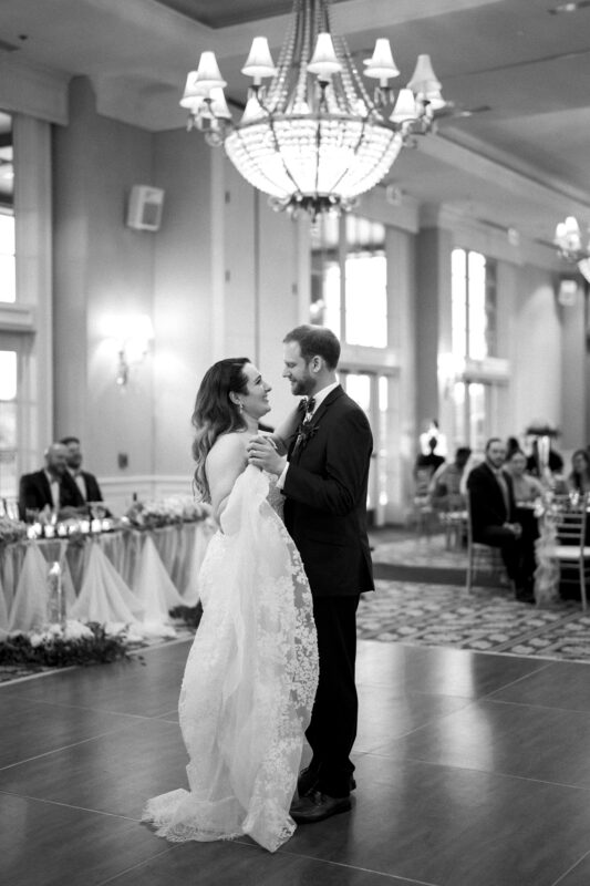 A bride and groom share their first dance at a river landing wedding in a grand ballroom with elegant chandeliers. Guests are seated at decorated tables in the background. The bride wears a lace-trimmed gown, and both smile as they embrace on the wooden dance floor.