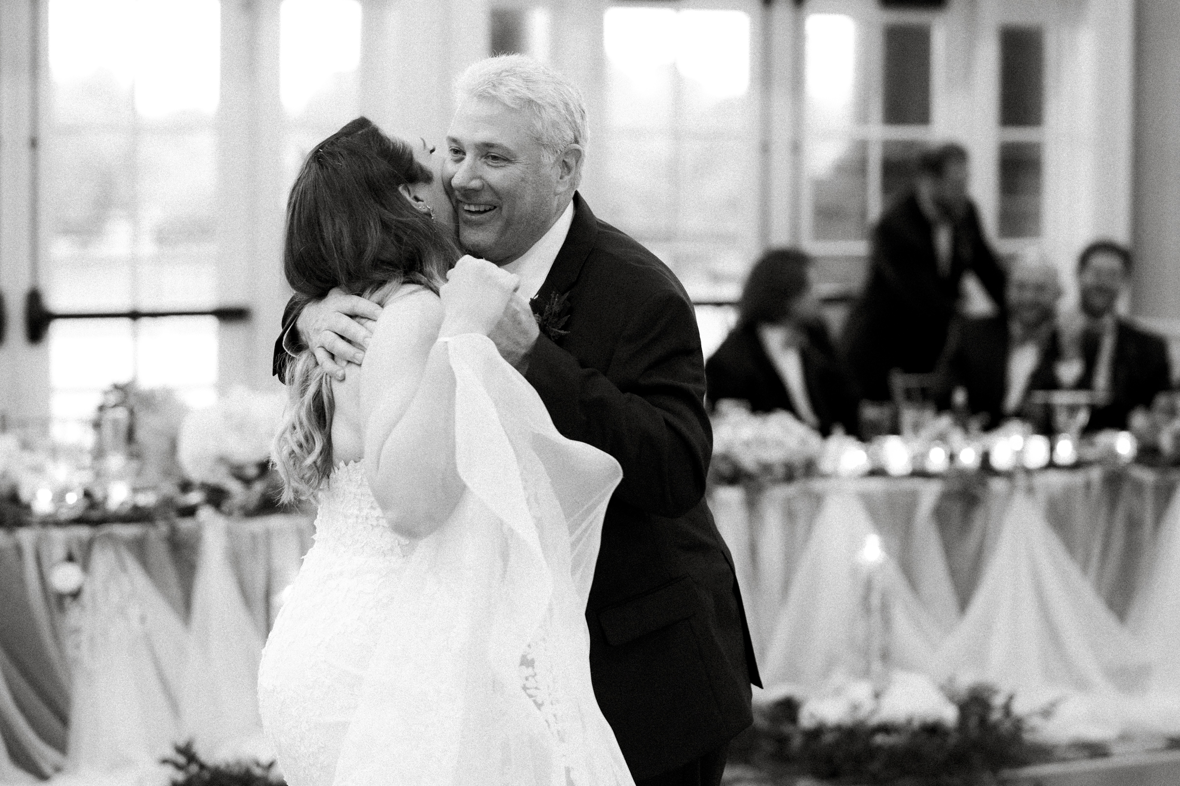 A black and white photo captures a joyful bride dancing with an older man, possibly her father, at the enchanting River Landing wedding reception. They're surrounded by a decorated table and guests in the background.