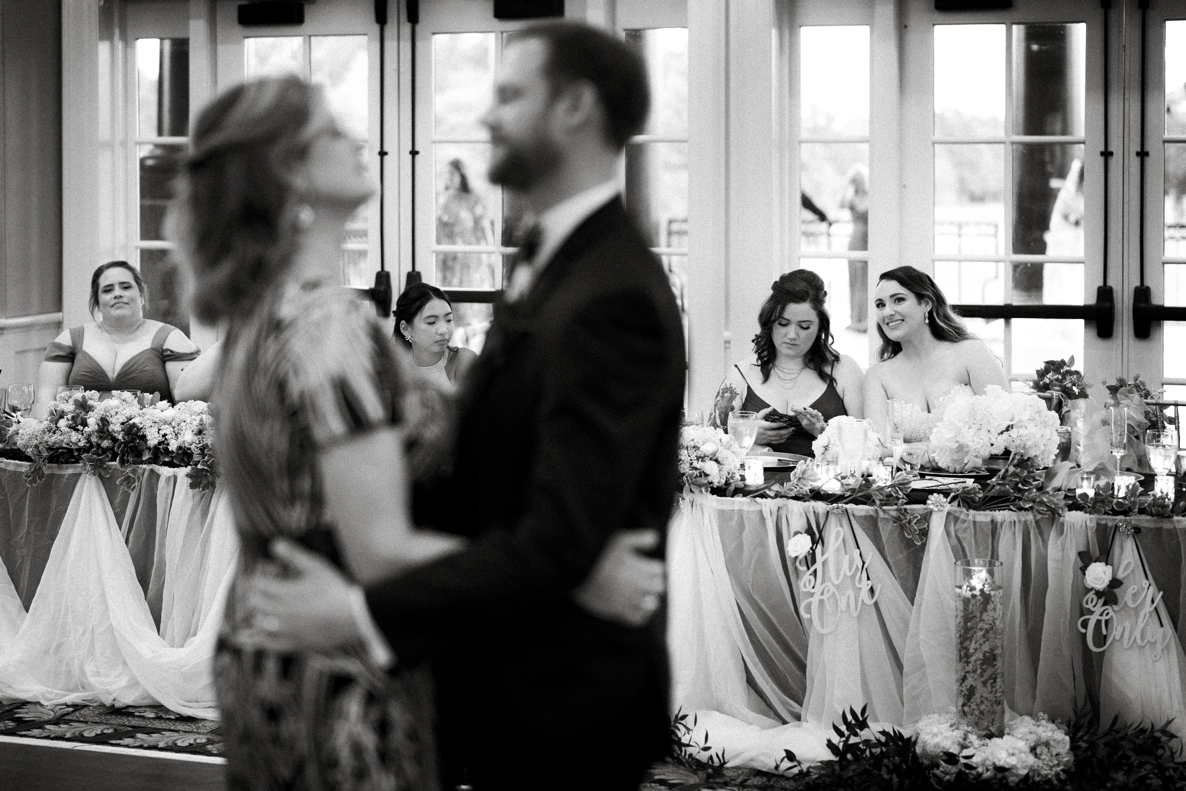 A couple dances in the foreground of a river landing wedding reception, slightly out of focus. In the background, a bridal party sits at a long table adorned with flowers. People at the table watch the dancing couple with smiles. Black and white image.