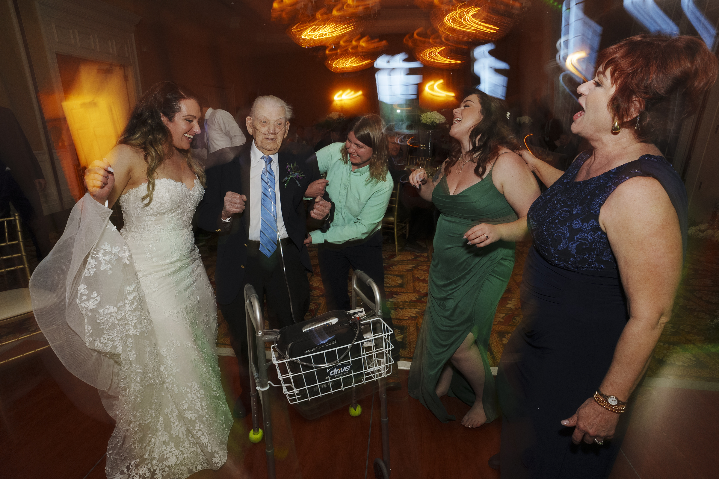 At a river landing wedding, a bride in a white gown and three women, two in green dresses and one in dark blue, joyfully dance with an elderly man using a walker. They celebrate amidst blurred lights and festive decor at the vibrant reception.