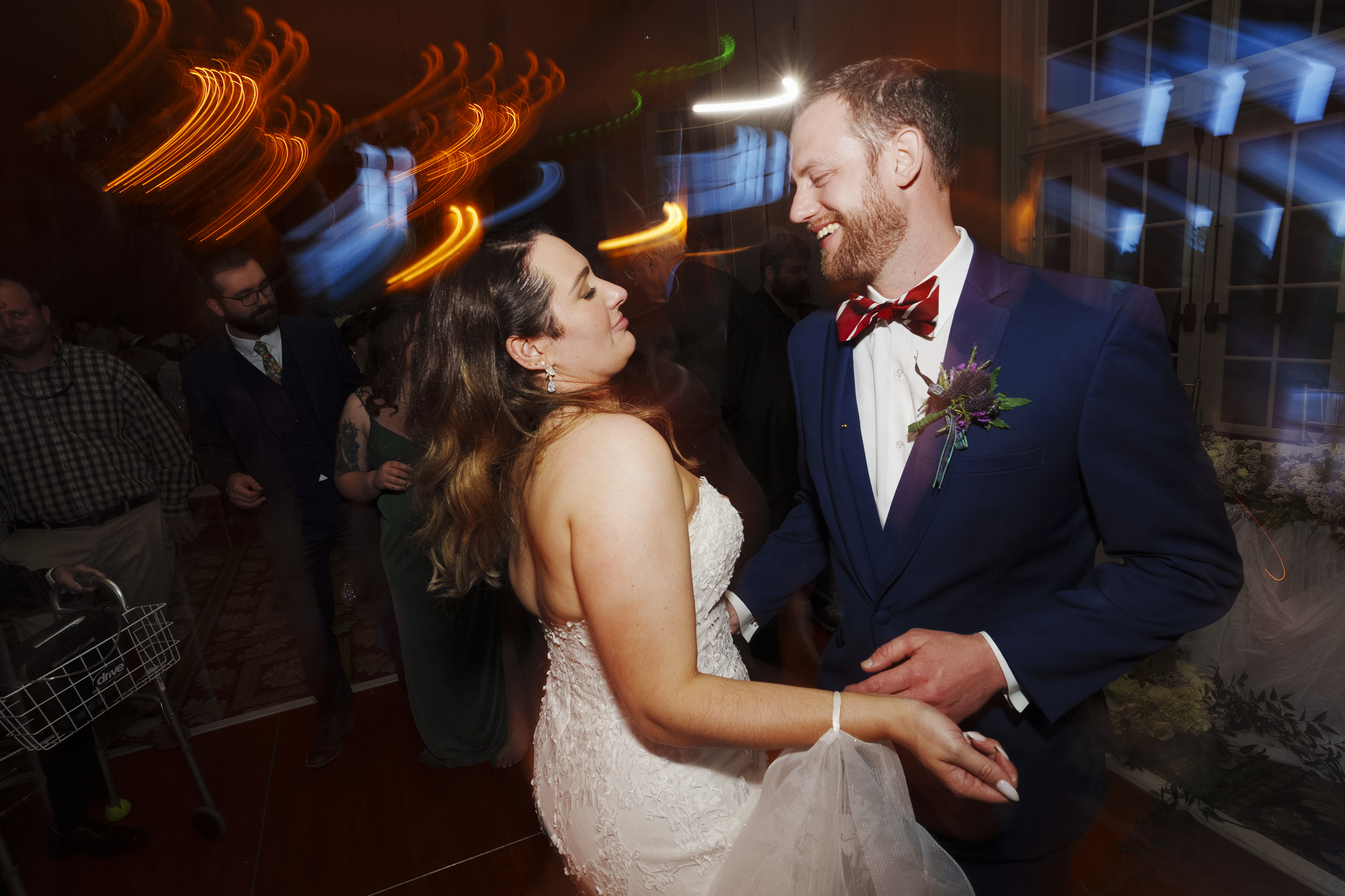 A bride and groom smile and dance closely at their River Landing wedding reception. The groom wears a blue suit with a red bow tie, while the bride dazzles in a white dress. Guests and colorful light trails create a vibrant backdrop, enhancing the evening's joy.