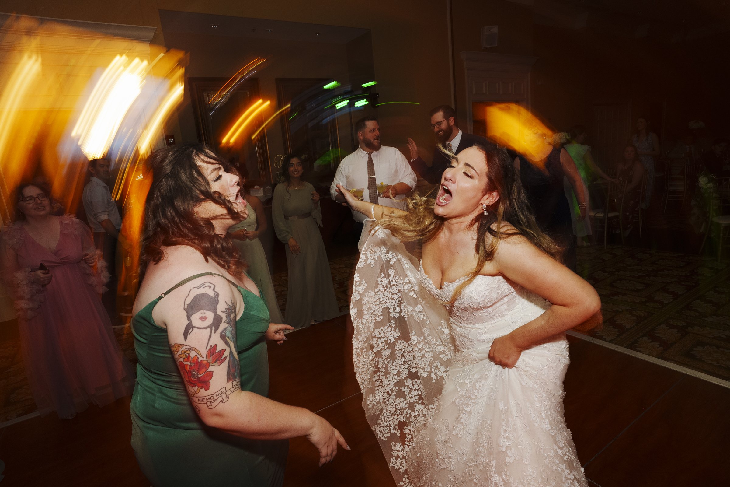 Two women joyfully dance at a lively River Landing wedding reception. One wears a white bridal gown, holding her dress up, while the other, with tattoos on her arm, dons a green dress. Guests in the background join in the celebration, with lights creating motion streaks.