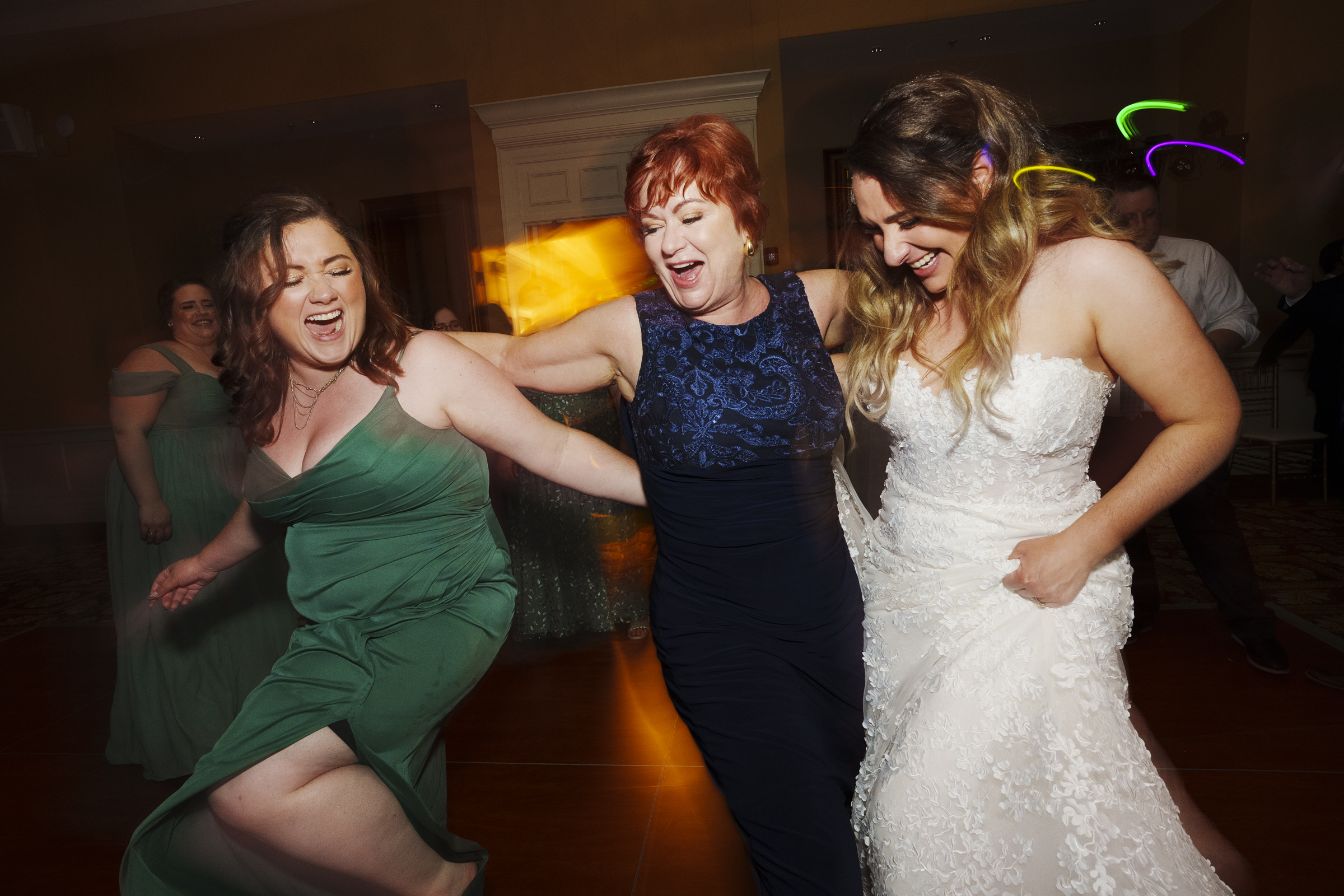 Three women joyfully dance together at a river landing wedding. One wears a green dress, another a blue dress, and the third a white wedding gown. They smile and embrace each other with lively expressions as colorful lights create a festive atmosphere.