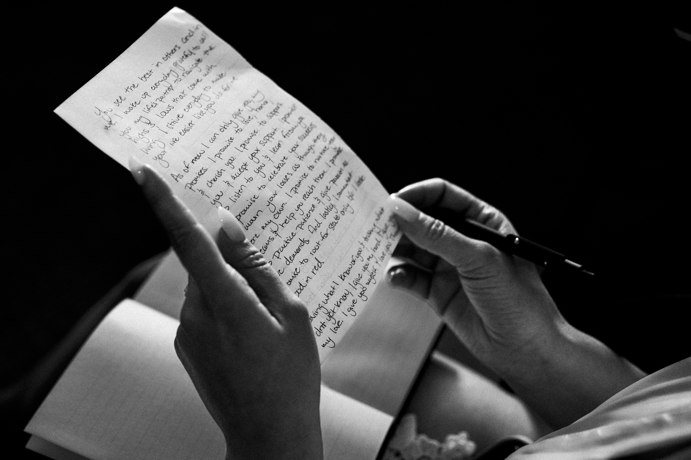 A person holds a handwritten letter and a pen, with a notebook resting on their lap, reminiscent of wedding vows crafted by the river landing. The black-and-white image focuses on the hands and paper, crafting an intimate and reflective atmosphere.