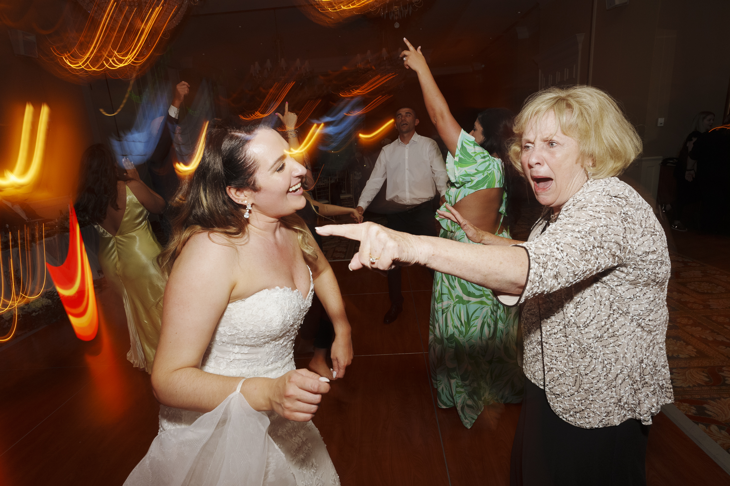 At the lively river landing wedding, a bride in a white dress dances energetically with an older woman in a floral blouse. The room buzzes with colorful motion blur, perfectly capturing the festive atmosphere as other guests join in on the dance floor.