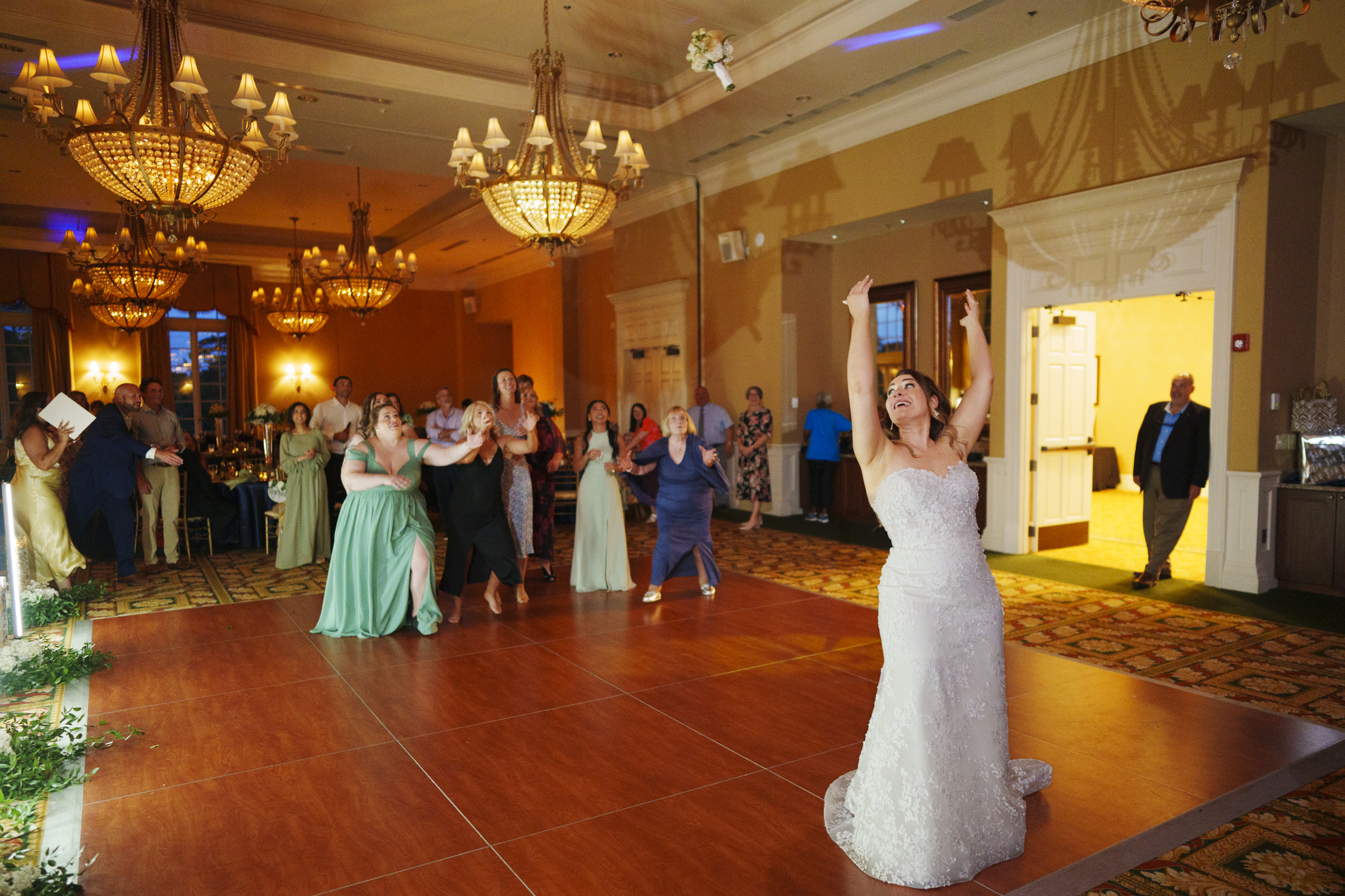 In an elegant river landing ballroom, a bride in a white dress prepares to throw her bouquet to the eager guests behind her. Chandeliers sparkle above as guests in formal attire eagerly await their chance to catch the special bouquet.