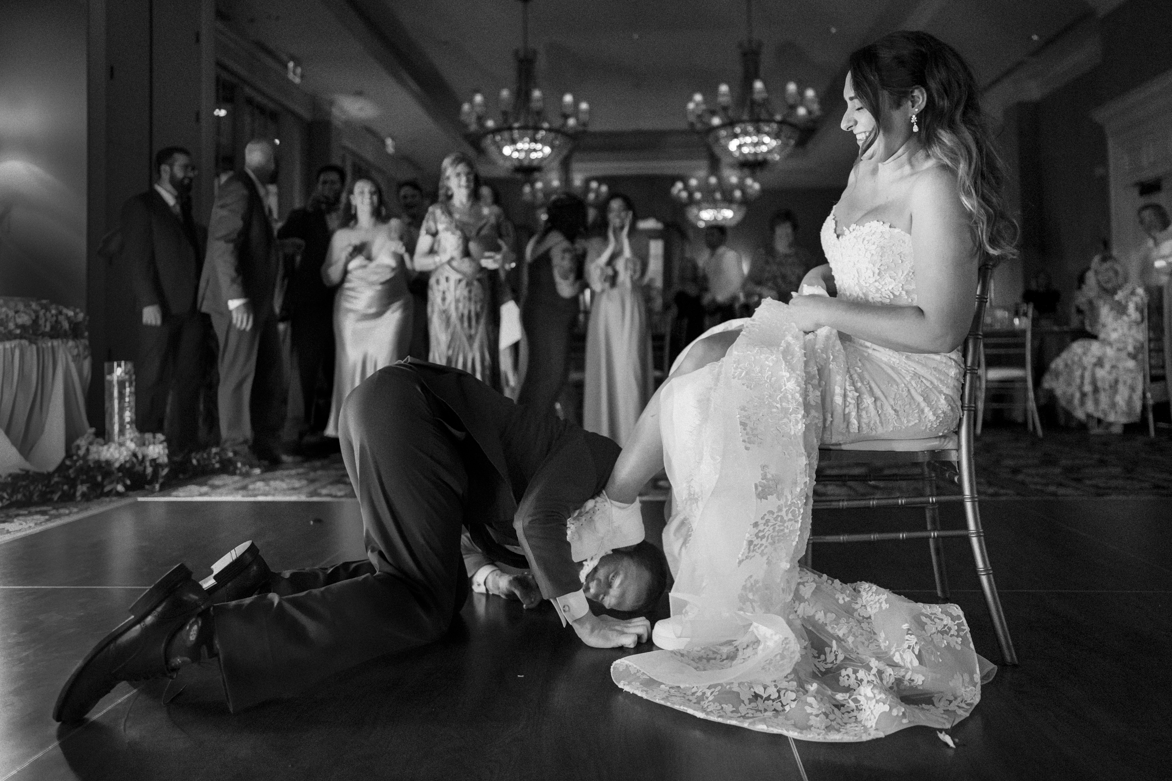 At a beautiful River Landing wedding, the groom kneels on the dance floor, playfully searching under the bride's dress for the garter during a fun ritual. The bride sits smiling on a chair as guests in formal attire watch and cheer in the background.