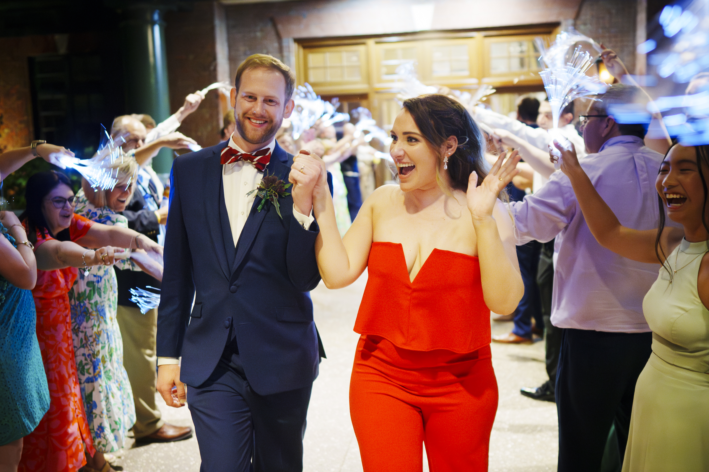 A couple joyfully walks through a tunnel of cheering guests waving sparklers at their river landing wedding. The man is in a blue suit and bow tie, while the woman wears a bright red outfit, adding to the festive atmosphere of this enchanting celebration.