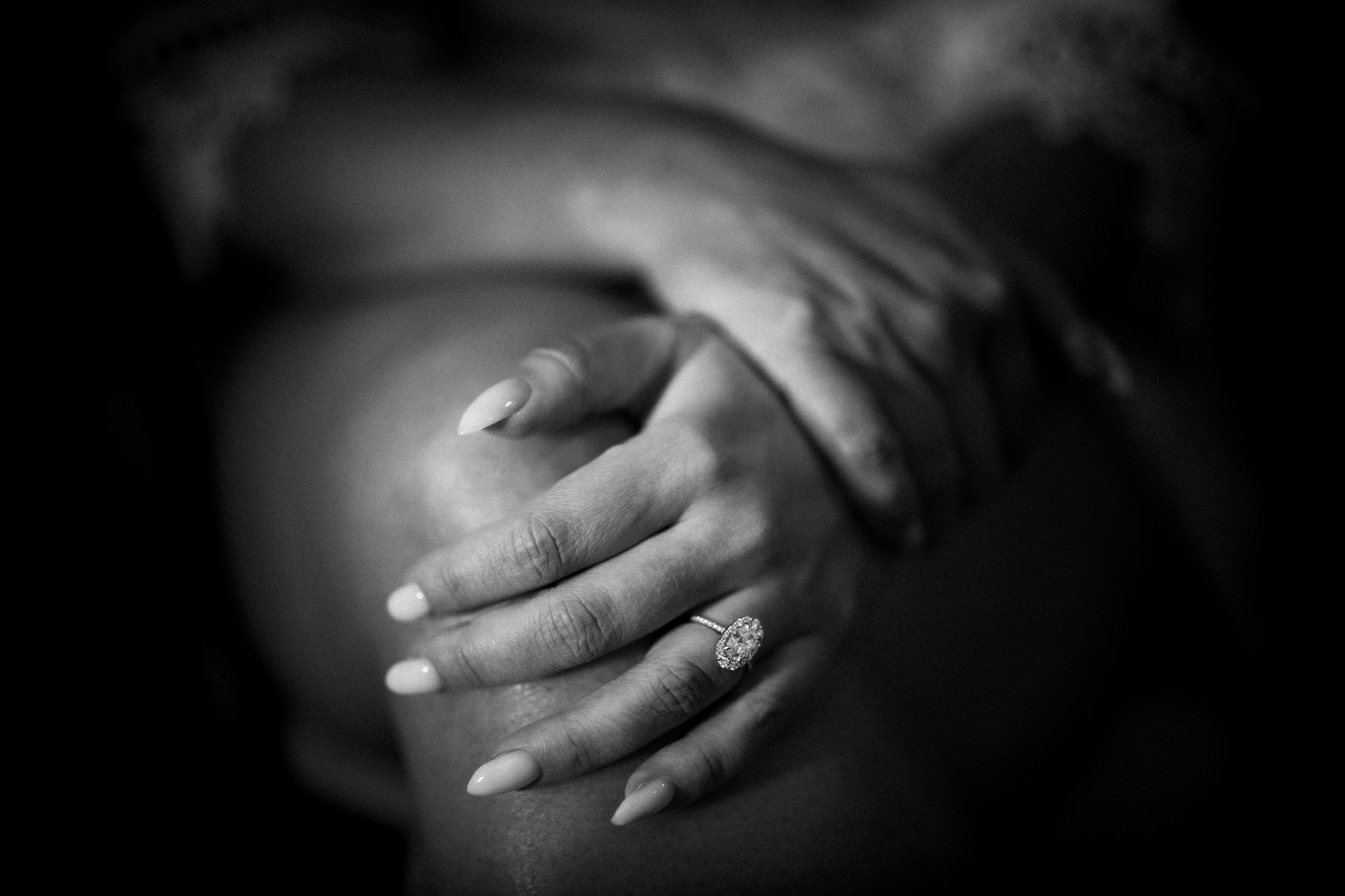 Black and white photo of hands resting on a knee, featuring long, manicured nails and a diamond engagement ring. The soft lighting evokes a contemplative mood, reminiscent of the serene ambiance at river landing weddings.