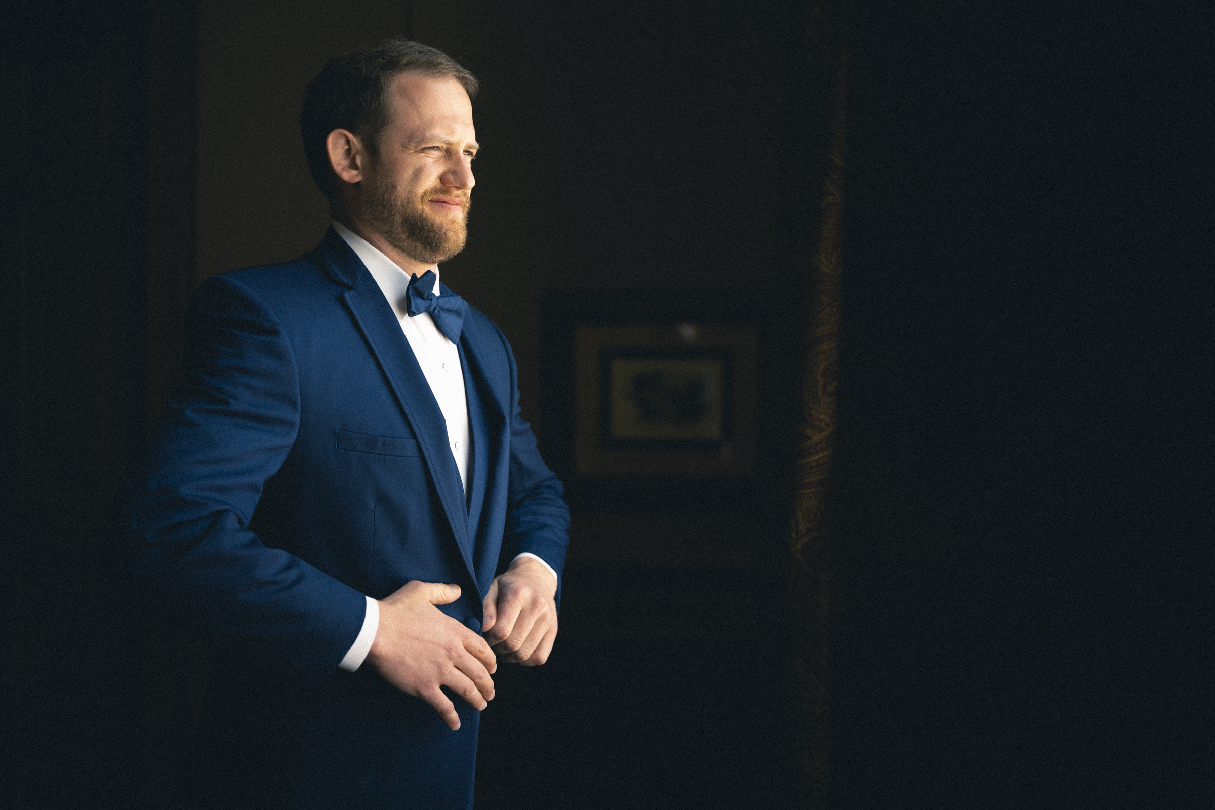 A man in a blue suit and bowtie stands in a dimly lit room, buttoning his jacket with practiced precision. He gazes out with a slight smile, illuminated by soft light from a nearby window, as if anticipating the joy of the river landing wedding. The background is dark, accented by a framed picture on the wall.