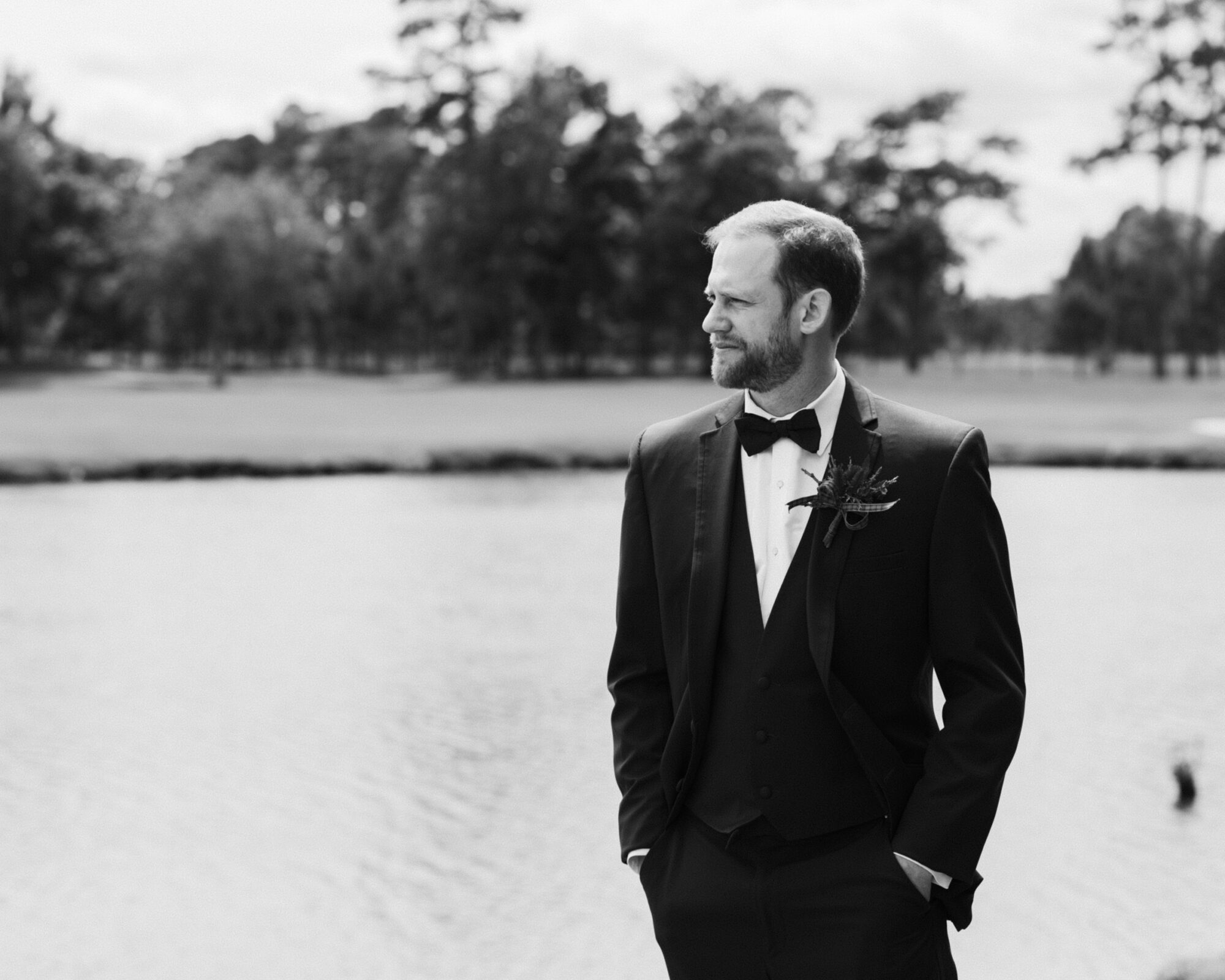 A man in a formal suit with a bow tie stands by the lake, hands in pockets, contemplating the serenity of the river landing. The background features trees and a calm body of water. The image is captured in elegant black and white, capturing wedding-like tranquility.