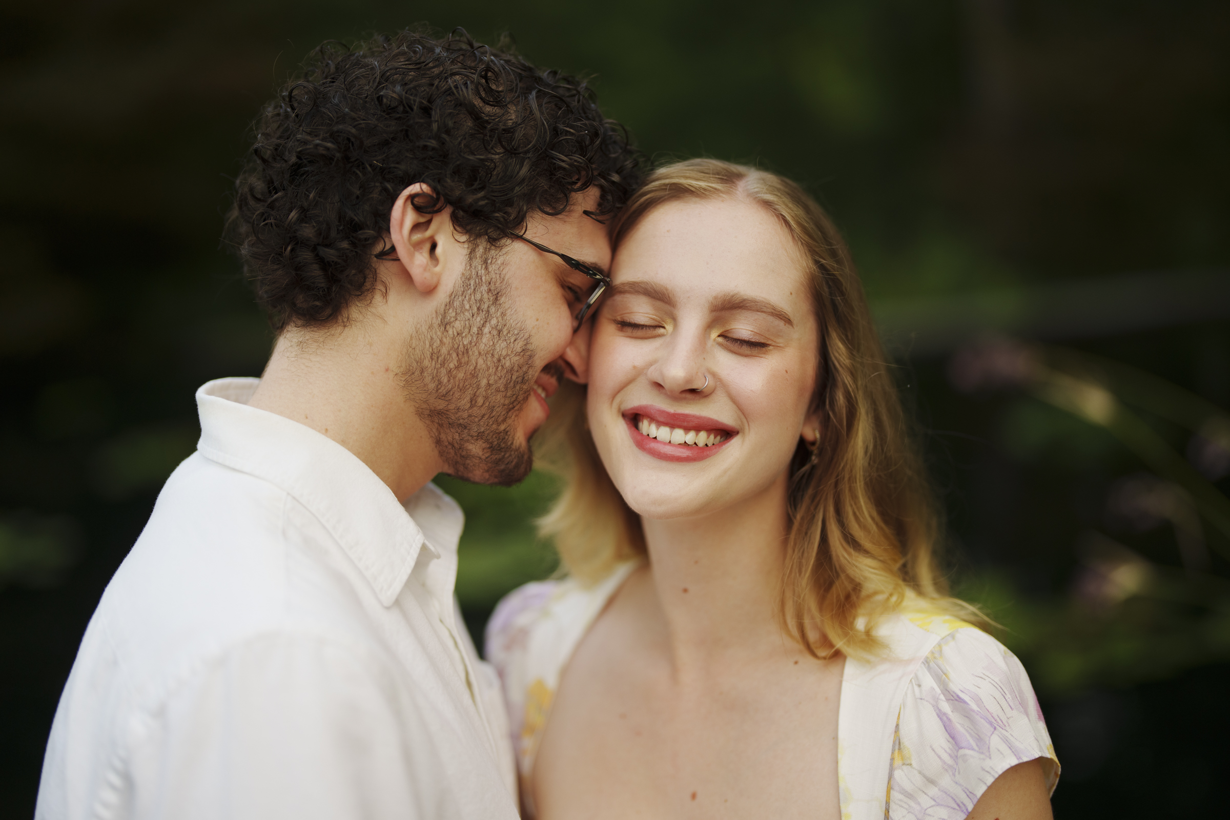 A couple stands closely together, smiling warmly during their Duke Gardens engagement session. The man, with curly hair and glasses, wears a white shirt, while the woman dons a floral dress. They appear content and joyful in this serene outdoor setting.