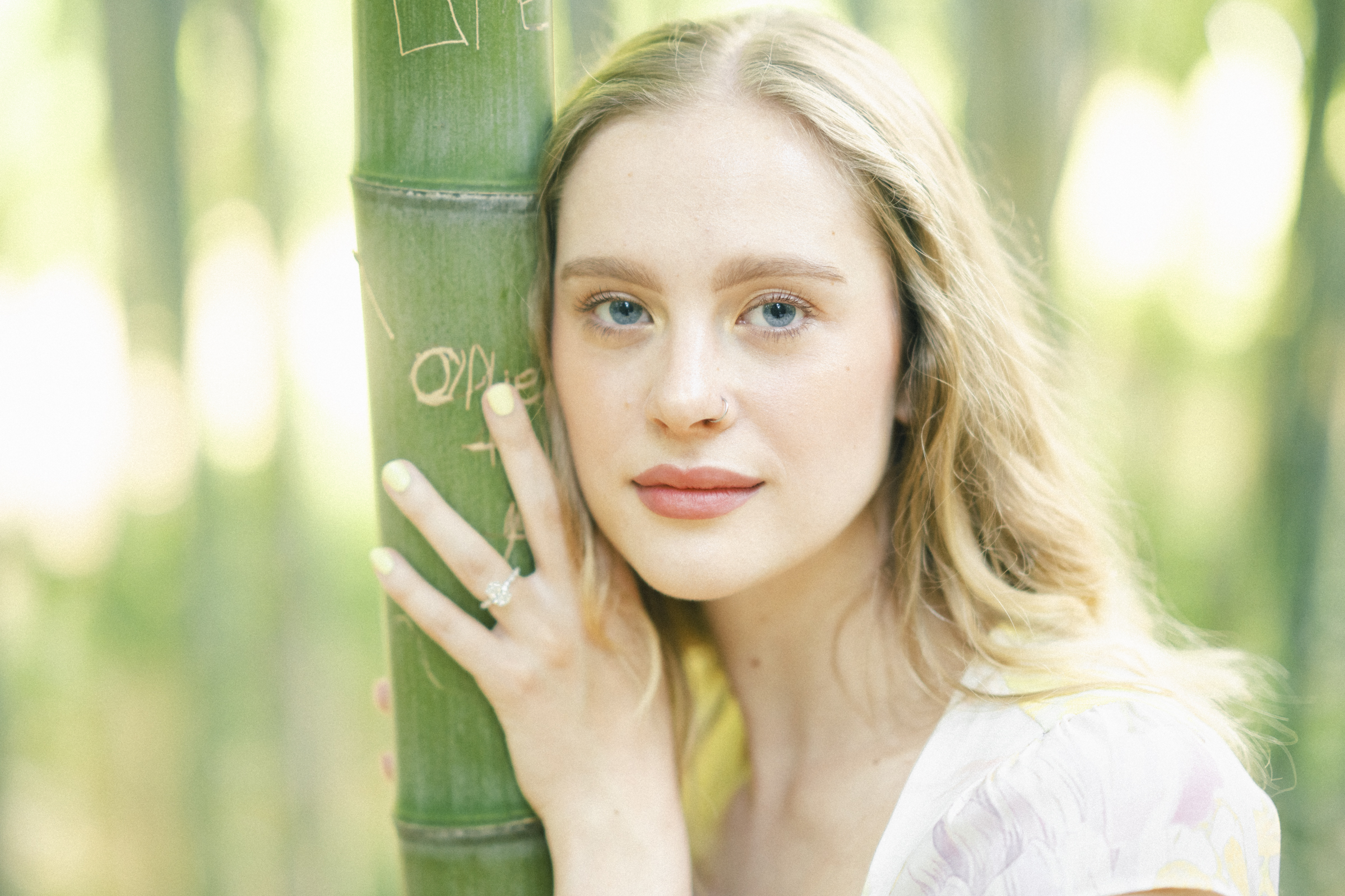 In a serene bamboo grove reminiscent of a Duke Gardens engagement session, a woman with long blonde hair gently rests her hand on a bamboo stalk. Clad in a white top, she exudes calm as soft natural light illuminates the scene.