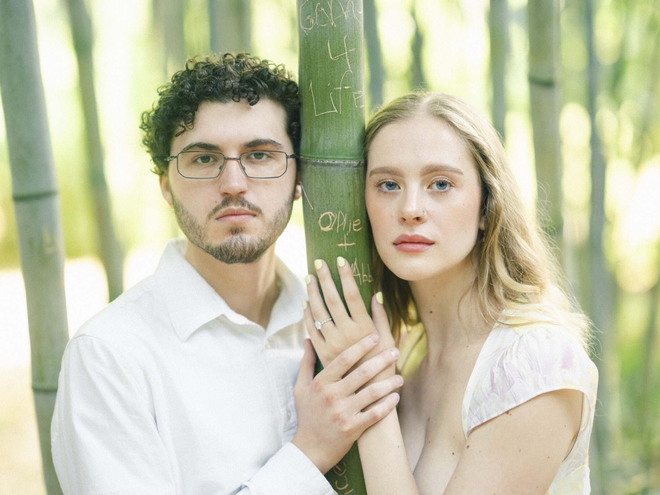 A young couple stands in a bamboo forest, capturing their engagement session's magic. The man, with curly hair and glasses, and the woman, with long blonde hair, wear light-colored clothing. Their hands gently touch the carved bamboo, reminiscent of Duke Gardens' serene beauty.