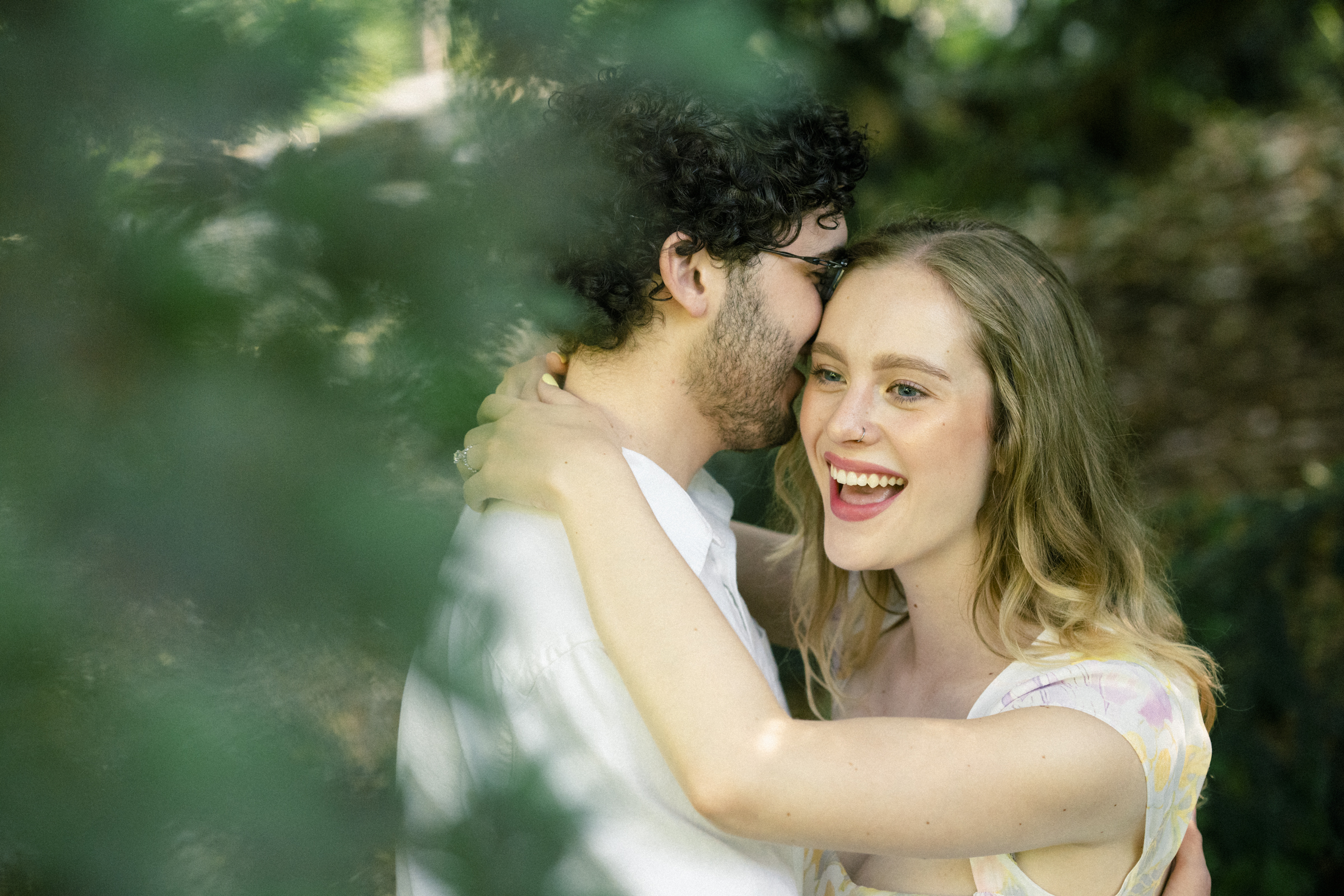 A woman smiling broadly embraces a man with curly hair in a sunlit, leafy setting at Duke Gardens. Her arms wrap around his neck as they share a joyful, intimate moment surrounded by greenery during their engagement session.