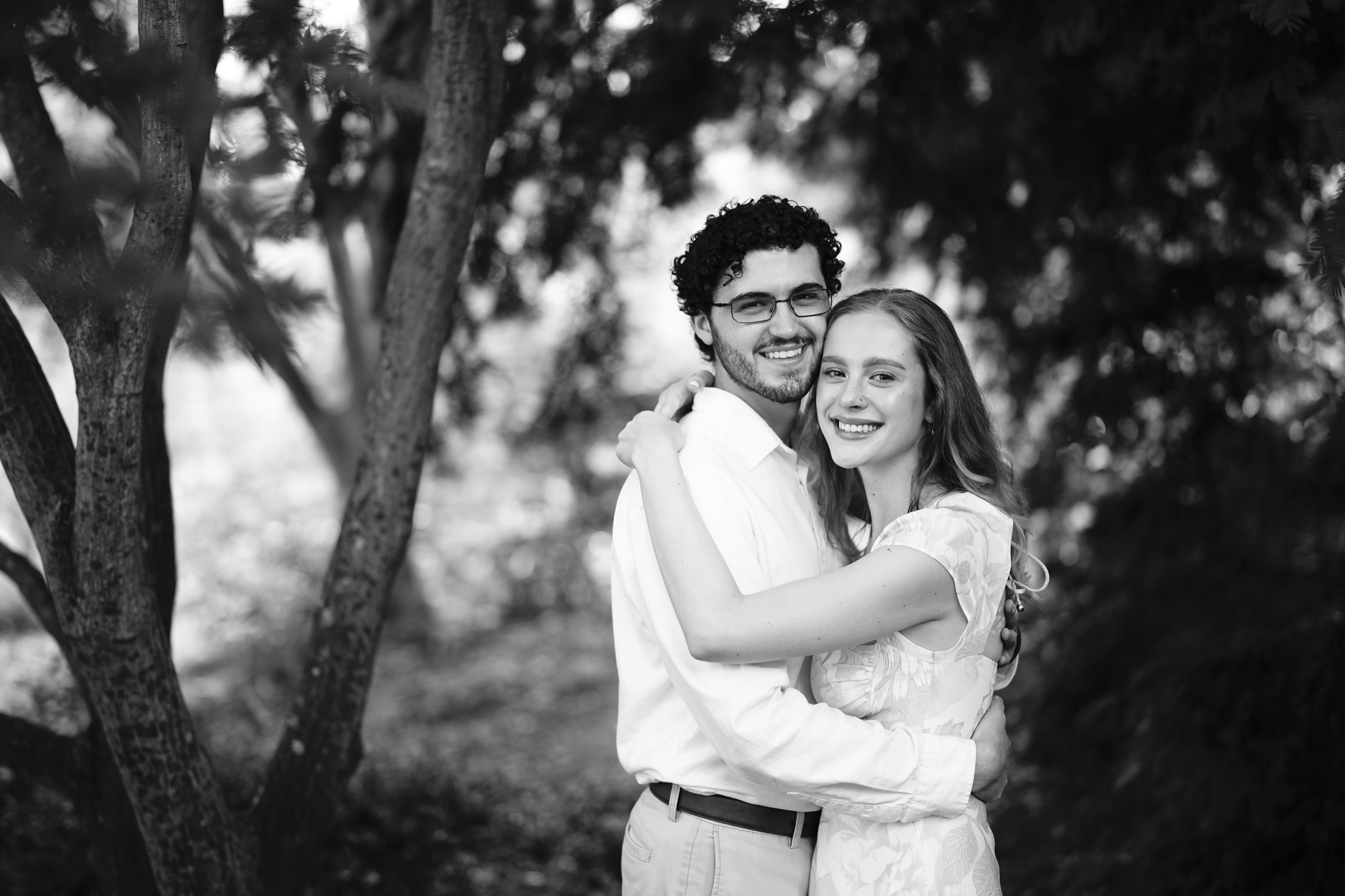 A black and white photo of a couple embracing outdoors during their Duke Gardens engagement session. They are both smiling and looking at the camera, with trees and foliage forming a natural, serene background.