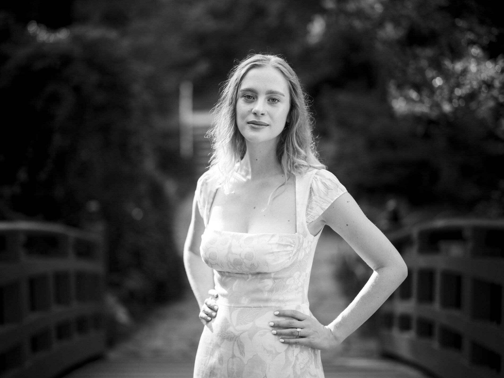 A woman in a floral dress stands confidently on a bridge at Duke Gardens, hands on her hips. The background is softly blurred, creating a serene, natural setting—perfect for an engagement session. The image is in black and white.