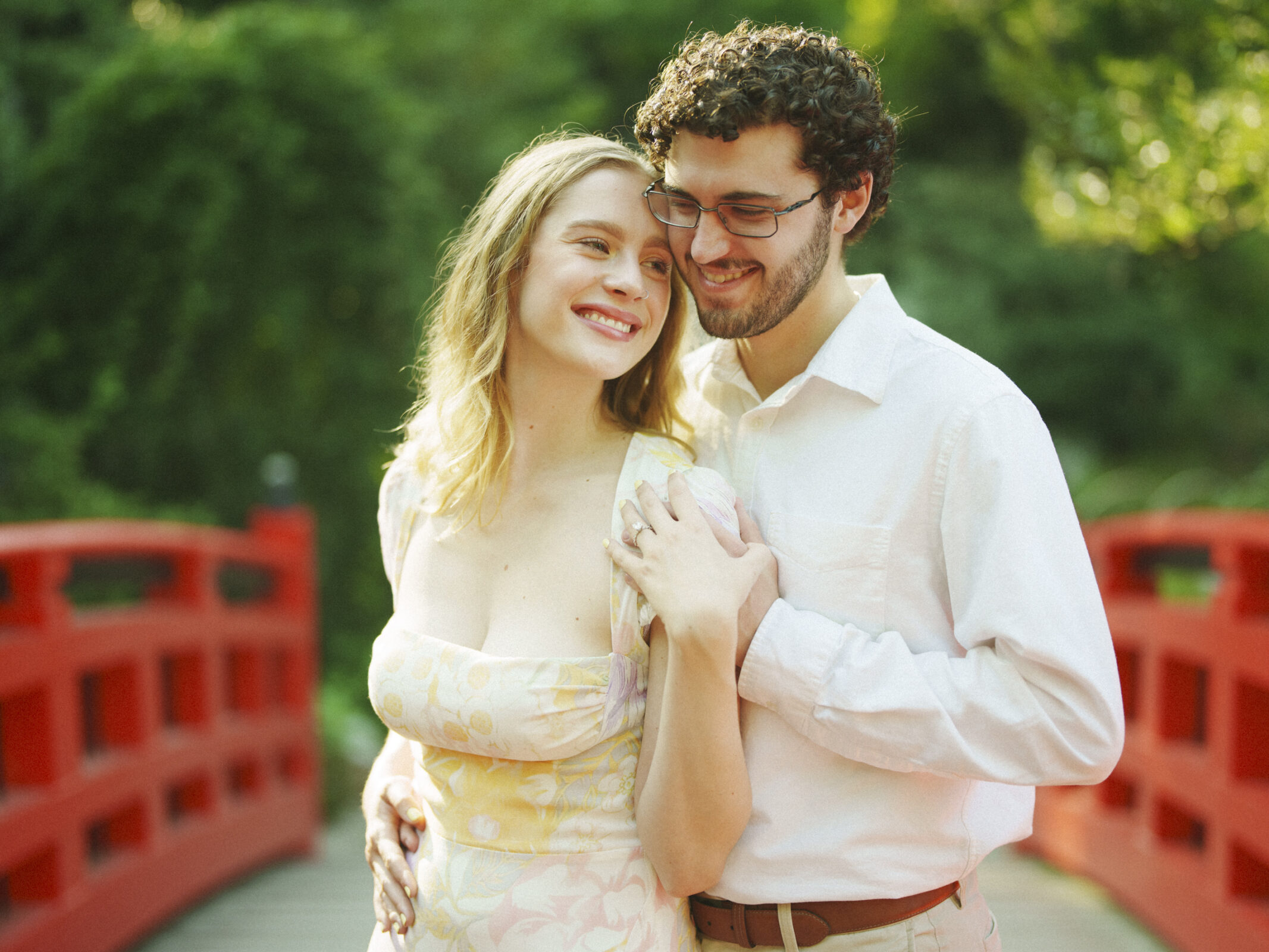 A couple stands on a red bridge in a lush garden during their Duke Gardens engagement session. The woman, in a light floral dress, smiles as the man, wearing glasses and a white shirt, gently embraces her. The scene is bathed in warm, natural light.