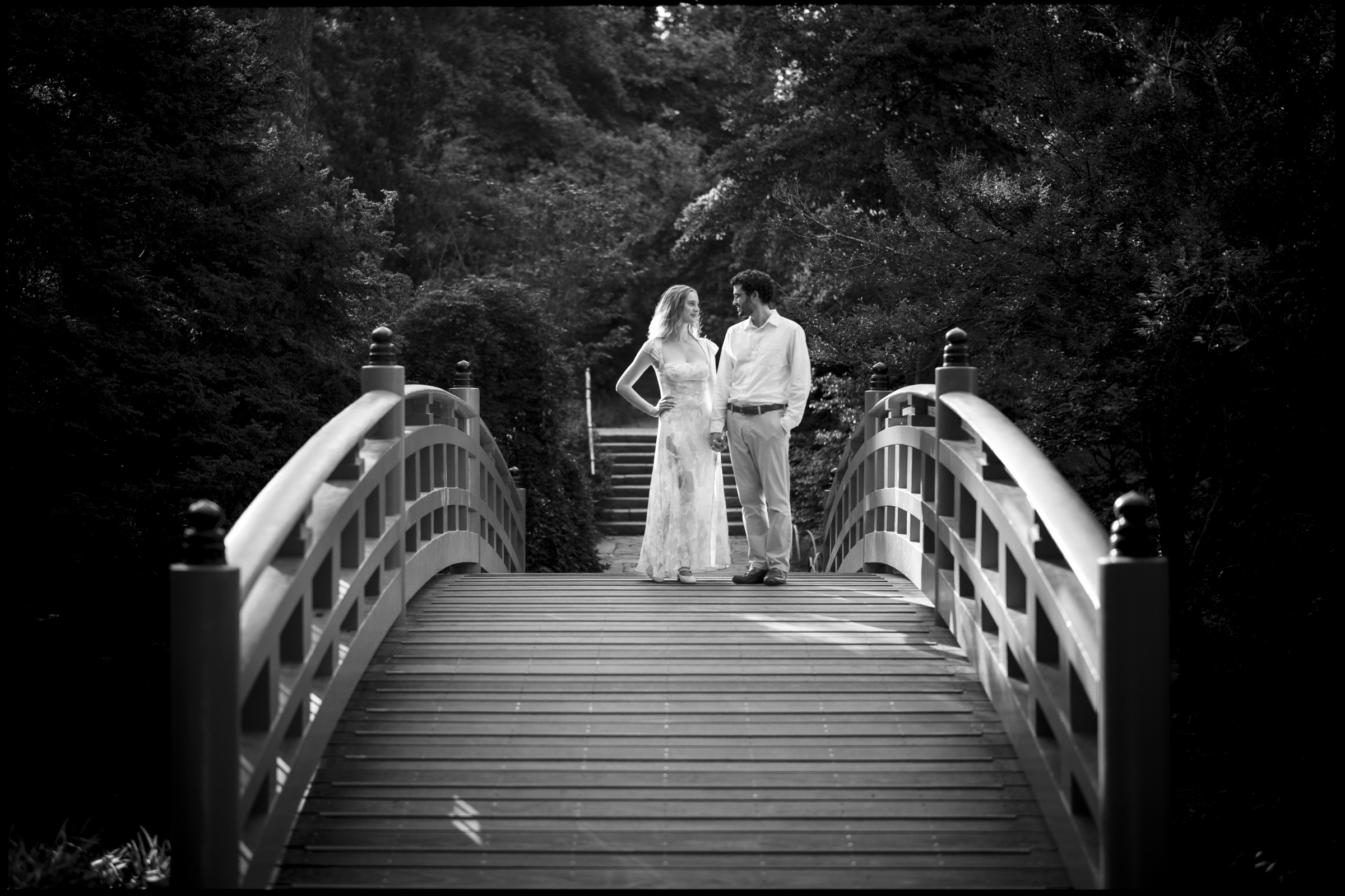 A couple stands on a wooden bridge in Duke Gardens, surrounded by lush trees. They are dressed in light, summery clothes and gaze at each other affectionately. The black-and-white image captures the timeless, romantic atmosphere of an engagement session.