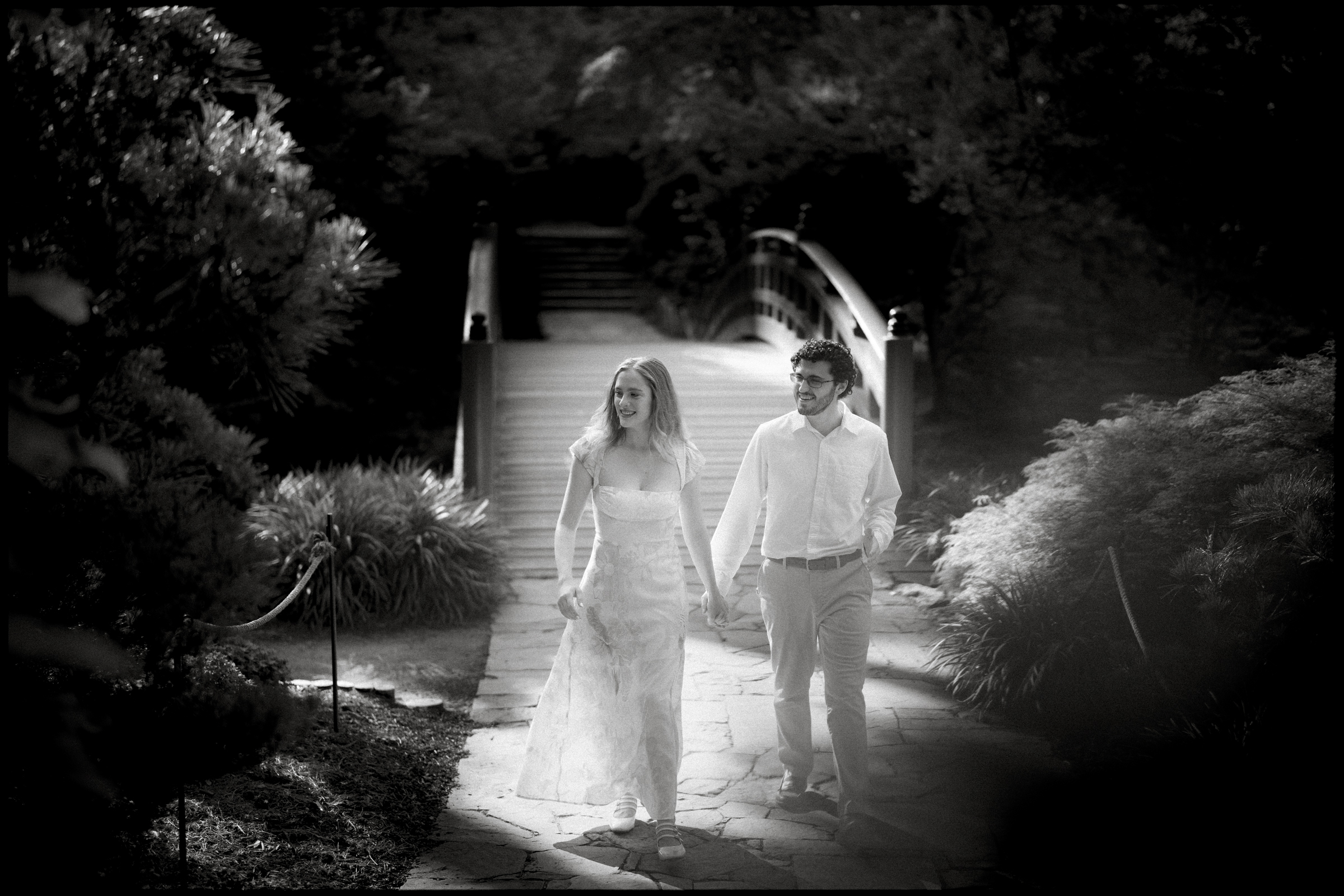 A couple walks hand in hand on a stone path through Duke Gardens, capturing the essence of their engagement session. Dressed in elegant attire, they approach an arched wooden bridge. The black and white scene highlights the serene and romantic atmosphere.