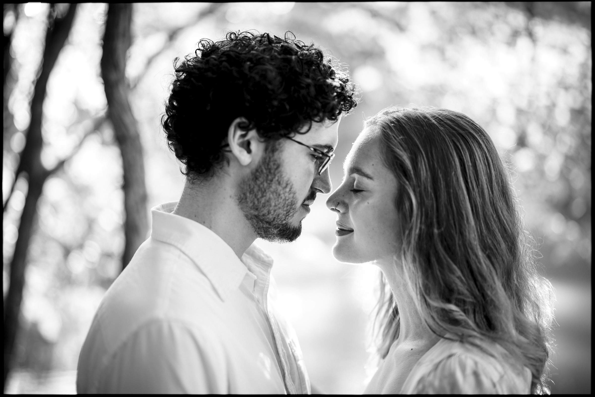 A black and white photo captures an intimate moment during a Duke Gardens engagement session. The couple stands closely, eyes closed, foreheads touching amidst the blurred backdrop of trees. Both are dressed in light-colored shirts, embracing the serene outdoor setting.