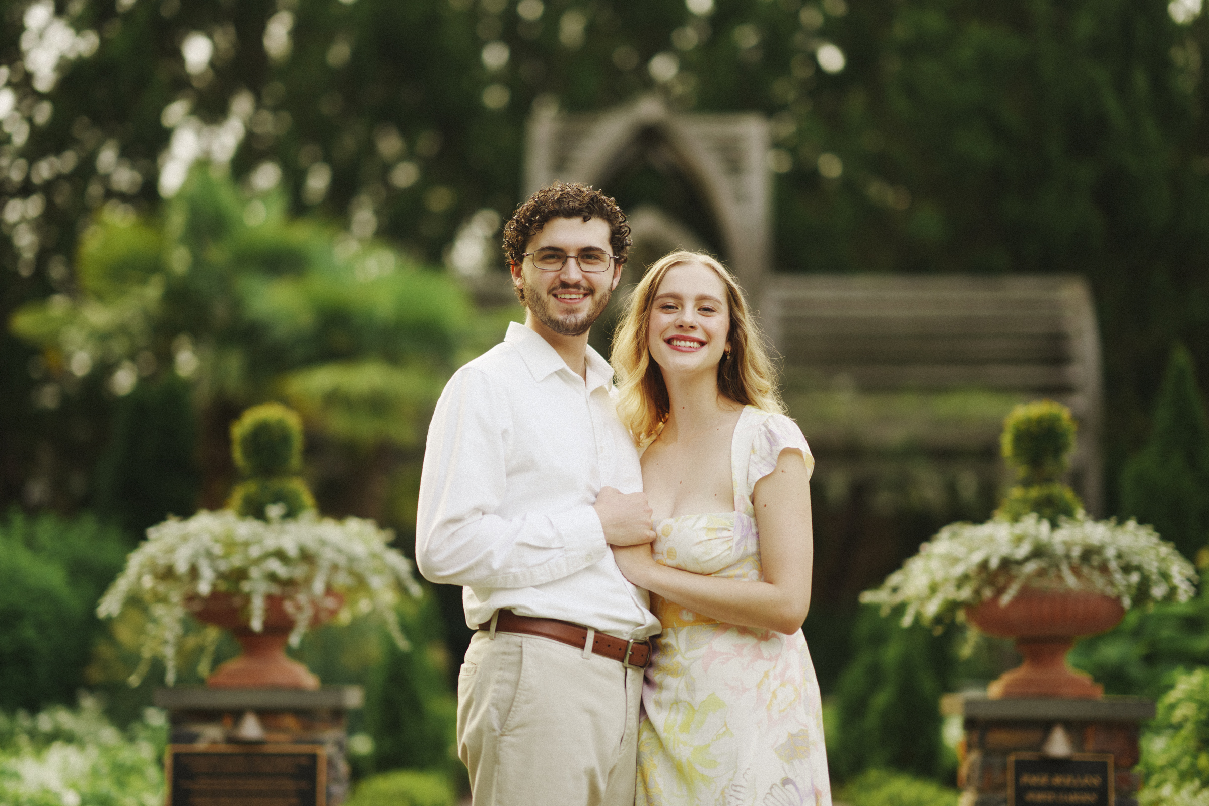 A smiling couple stands outdoors in the lush Duke Gardens, capturing a cherished engagement session. Both are dressed in light-colored clothing. The background features greenery and a wooden structure, enhancing the serene, romantic atmosphere.