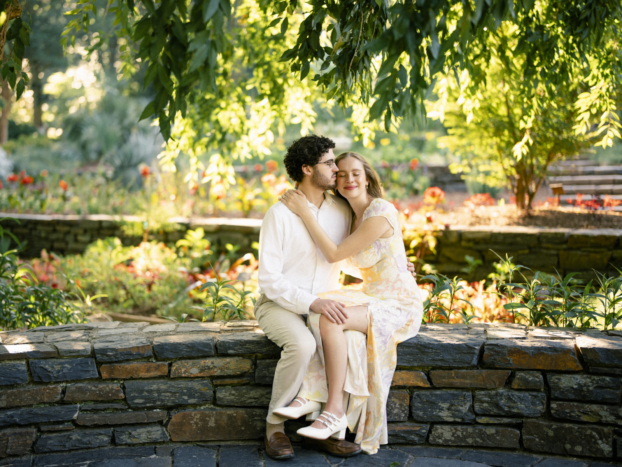 A couple sits on a stone wall in Duke Gardens, capturing their engagement session's essence. The woman, in a floral dress, embraces the man in a white shirt and beige pants. Sunlight filters through the leaves, casting a warm glow as they appear happy and relaxed.